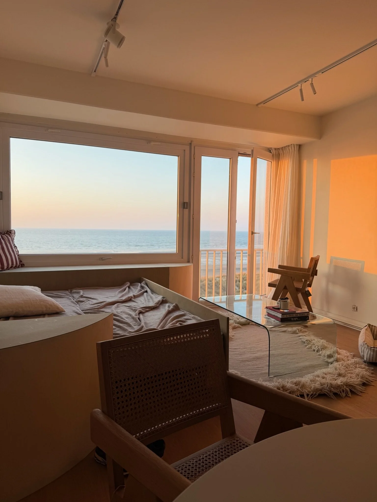A cozy bedroom with a large window taking up most of the wall, overlooking the ocean at sunset. The bed is unmade with beige bedding. A small table near the window holds books, a candle, and a decorative item. There are chairs, a fluffy rug, and curt