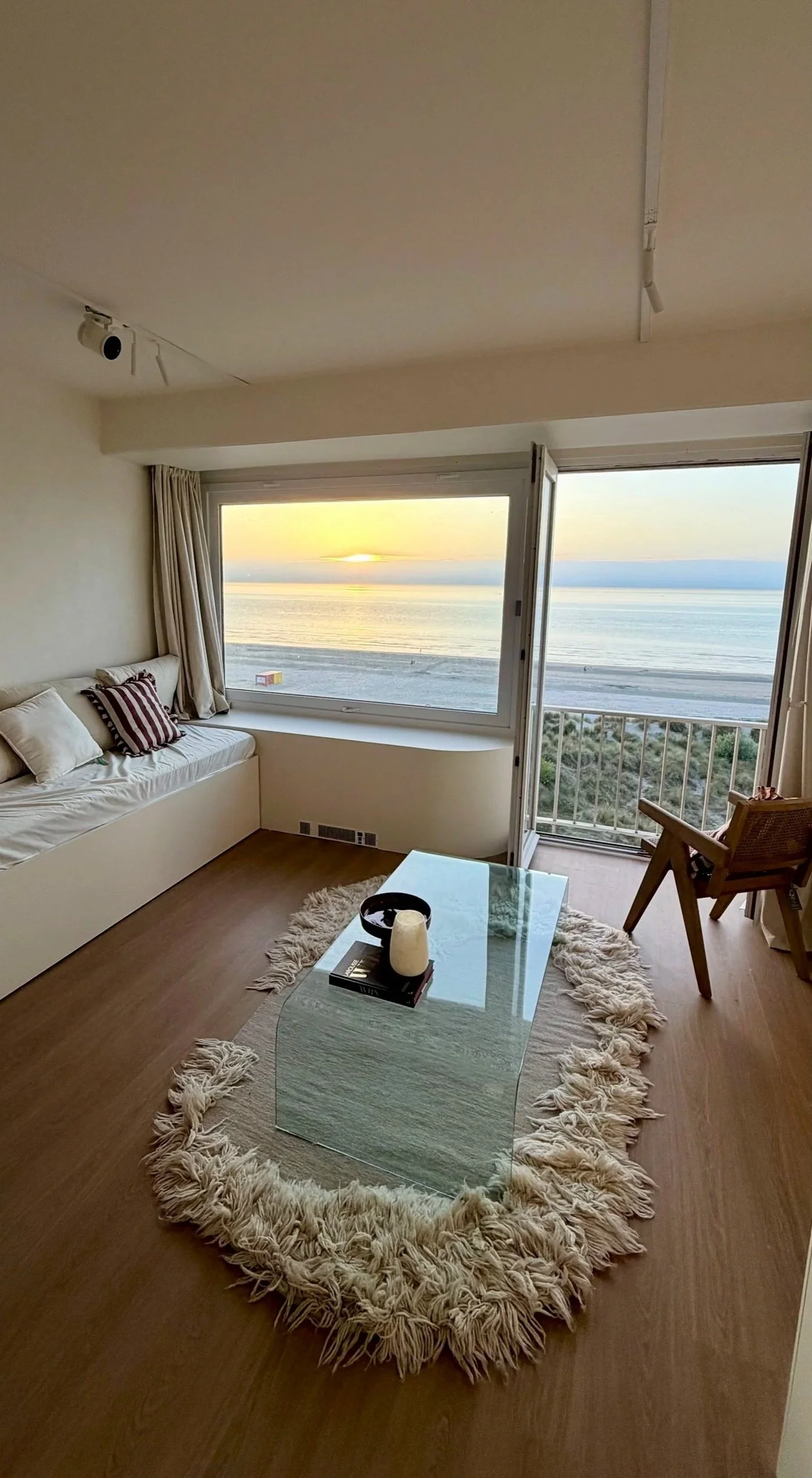 Living room with large window overlooking the ocean, beige sofa with striped pillows, glass coffee table on a shaggy rug, wooden chair, and coastal decor.