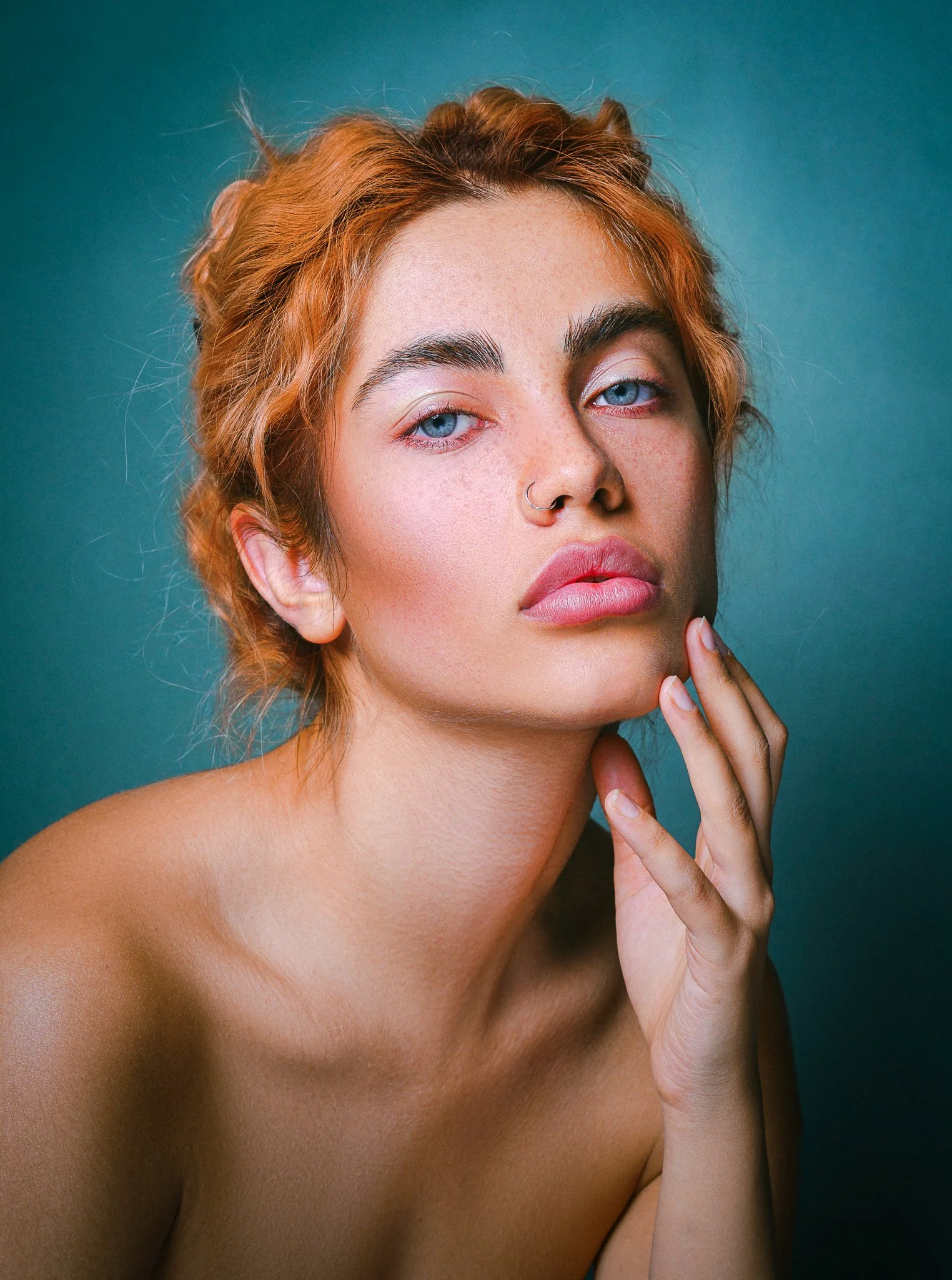 Porträt eines jungen Mädchens mit roten Locken, blauen Augen und Sommersprossen, leicht nackt mit Hand an Kinn, vor blauem Hintergrund.