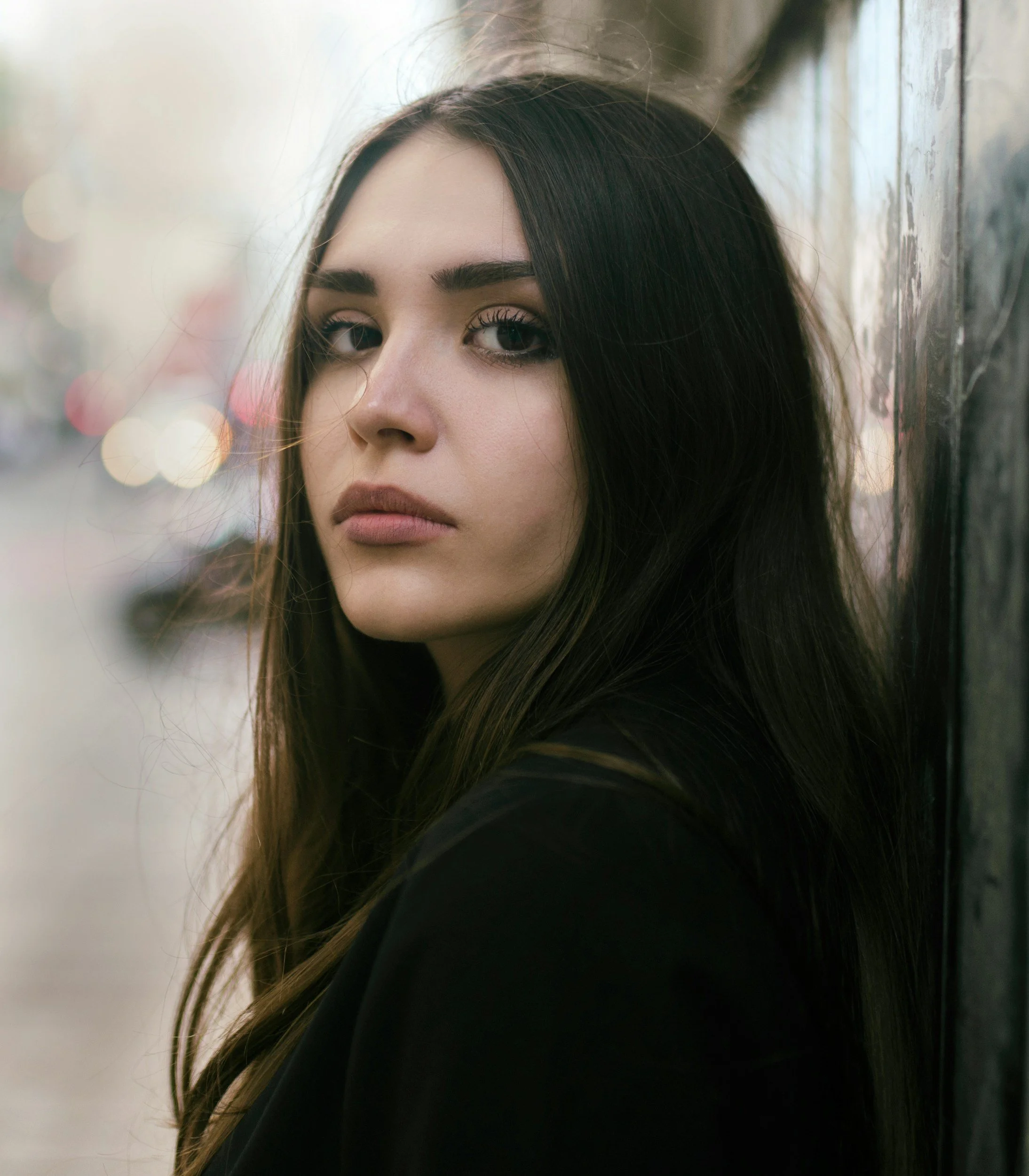 Nahaufnahme eines jungen Mädchens mit langen schwarzen Haaren, dunklen Augen und natürlichem Make-up, das gegen eine Holzverkleidung lehnt, im Hintergrund verschwommene Lichter und Stadtlichter.