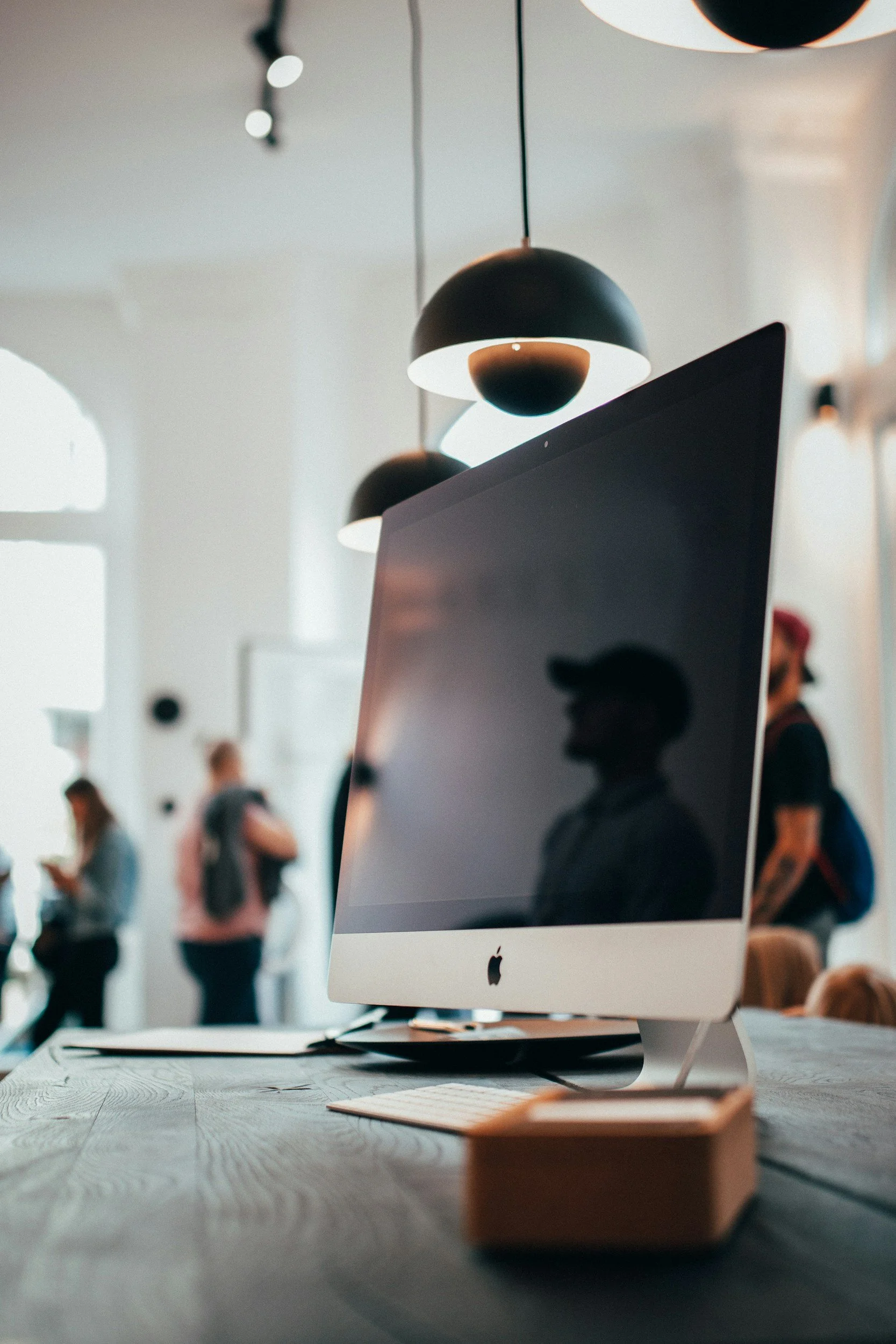 Ein heller Raum mit modernen schwarzen und weißen Designlampen. Auf einem Tisch steht ein Apple-Computer mit großem Bildschirm, im Vordergrund liegt ein kleiner Holzteller.