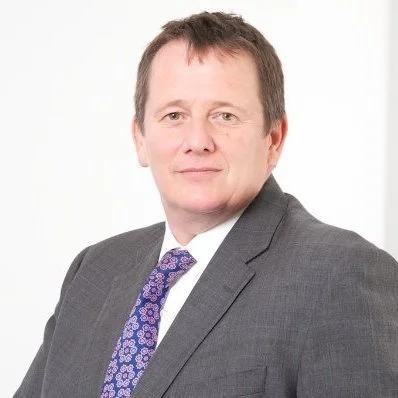 Professional headshot of a middle-aged man with short brown hair, wearing a gray suit, white shirt, and purple patterned tie, against a white background.