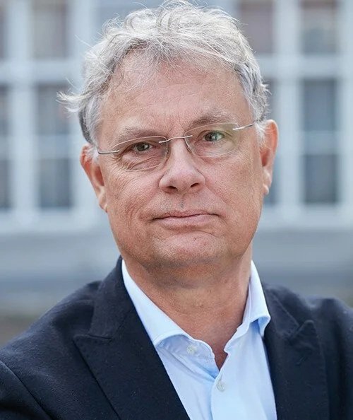 A portrait of an older man with gray hair, glasses, wearing a dark blazer over a light blue shirt, in front of a modern building with glass windows.
