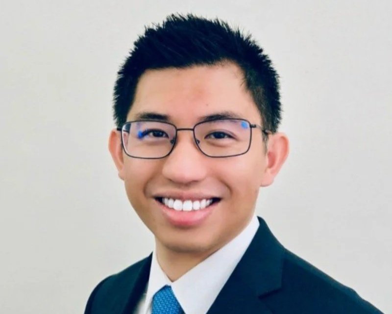 Young man with black hair, wearing glasses, a suit, and tie, smiling at the camera.