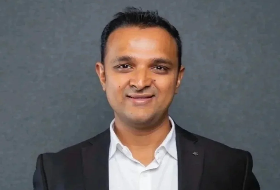 A man in a white shirt and black blazer smiling, standing against a gray background.
