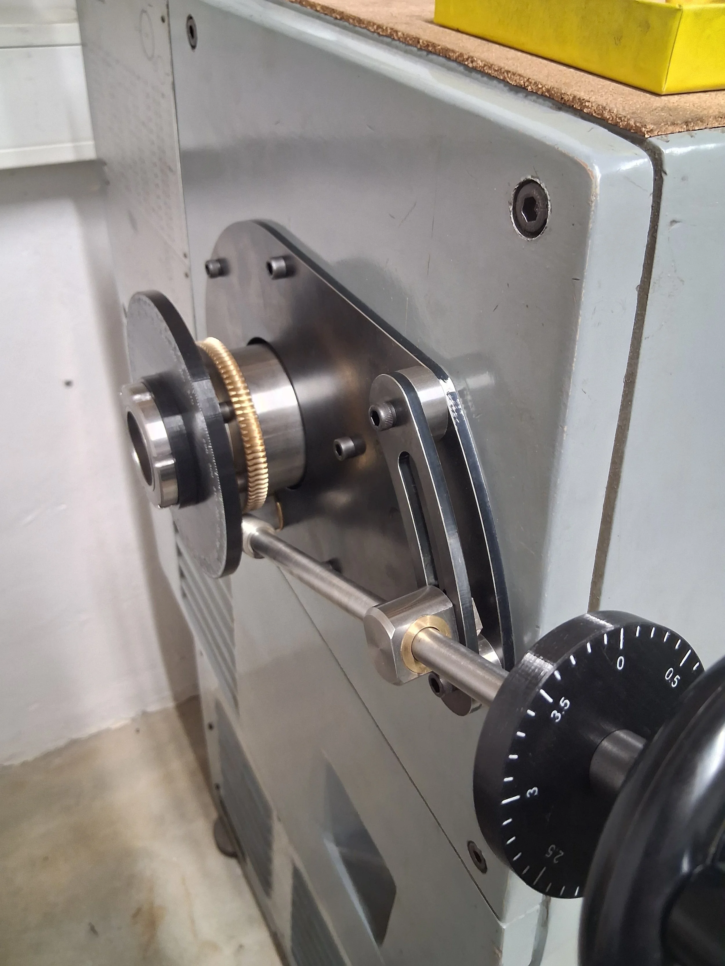 Close-up of a mechanical device with a scale and gear mechanism, attached to a gray metal surface with some visible screws and a yellow object on top.