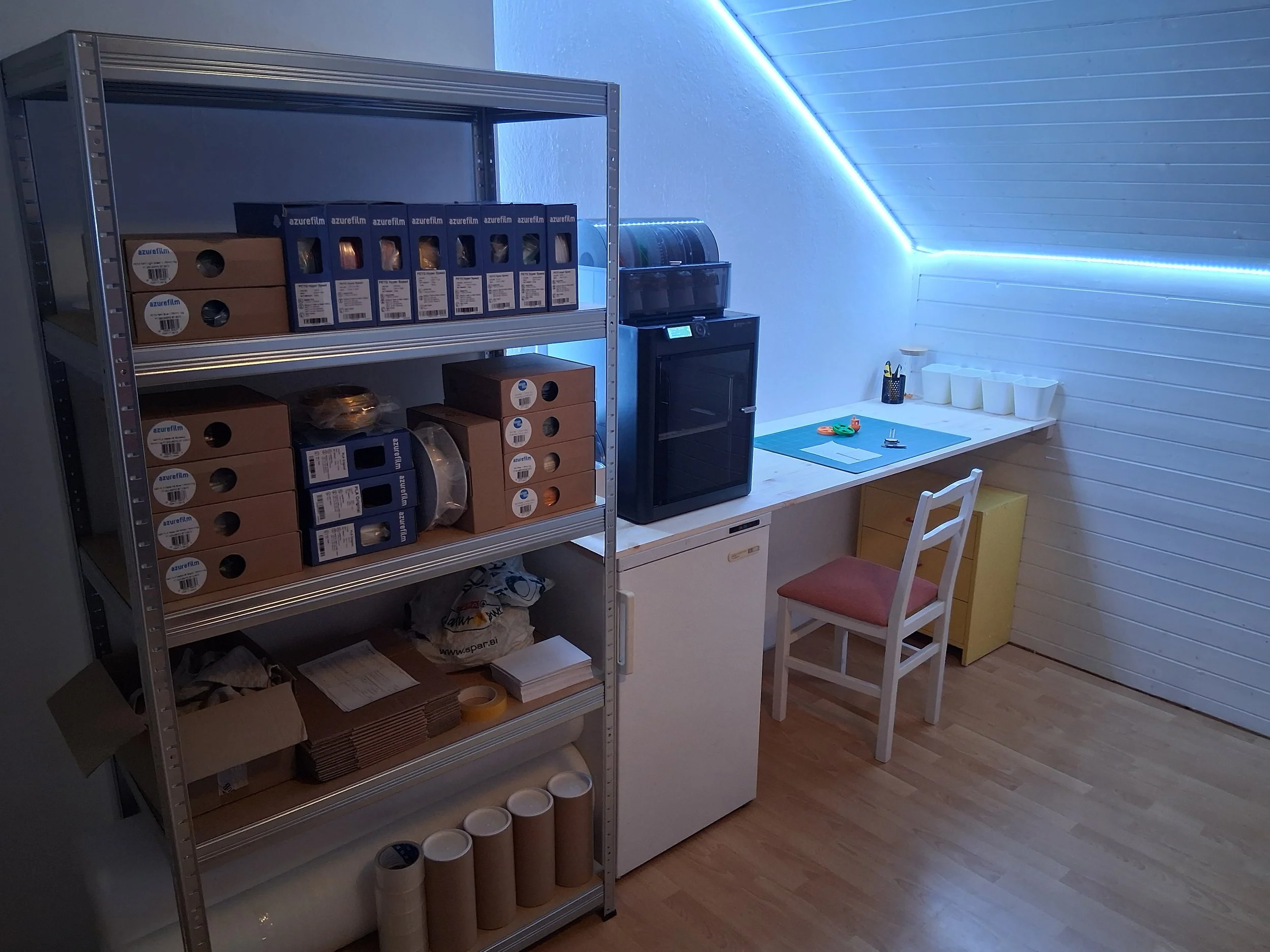 A room with a metal shelving unit filled with boxes and rolls of material, a small black oven, a counter with four white cups, a pen holder with colored pens, and a white chair, all under a slanted ceiling with blue LED strip lighting.