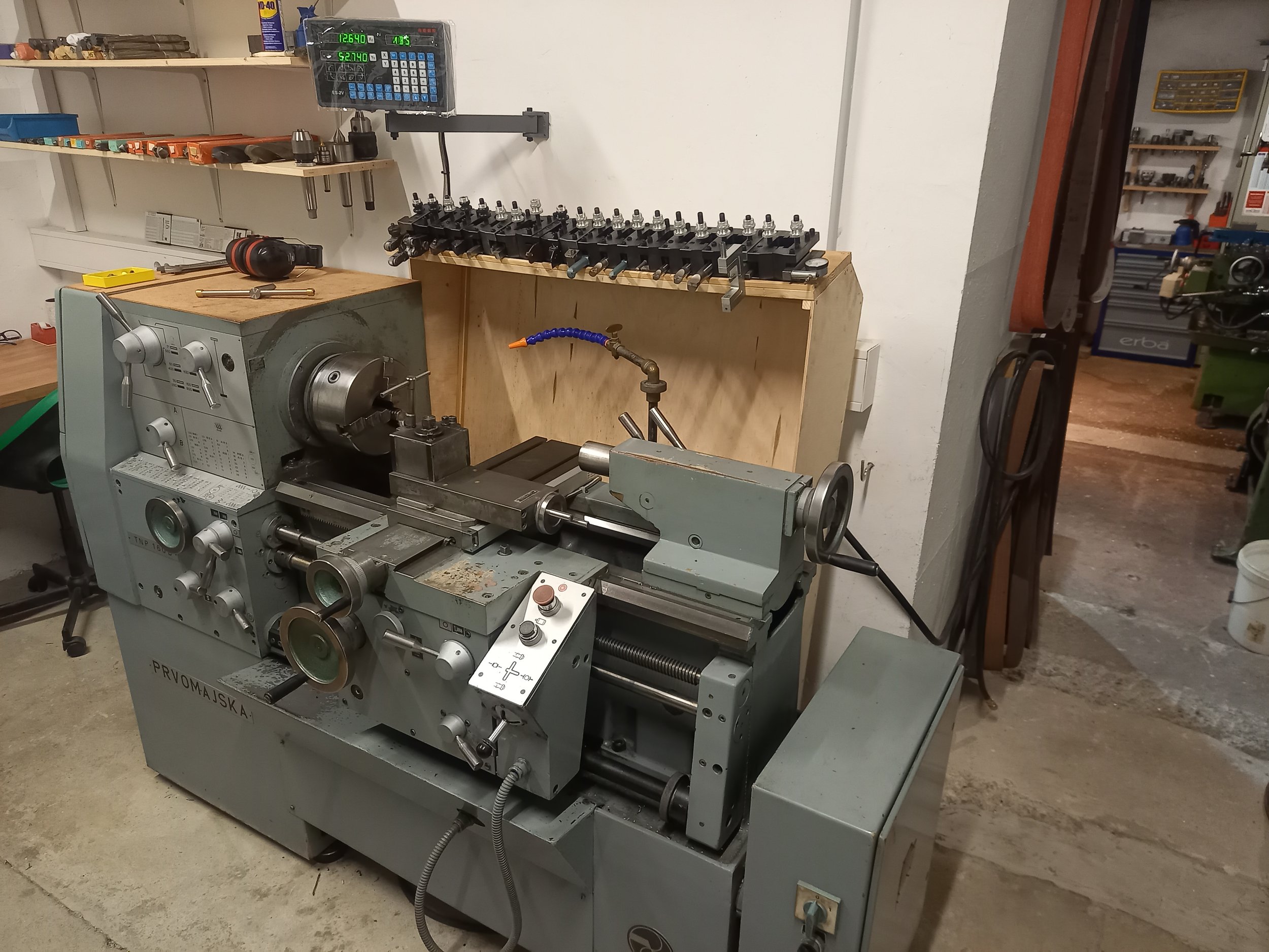 A workshop with a metal lathe machine in the foreground, and a workbench with various tools and supplies on the back wall. A digital control panel is mounted on the wall above the workbench.