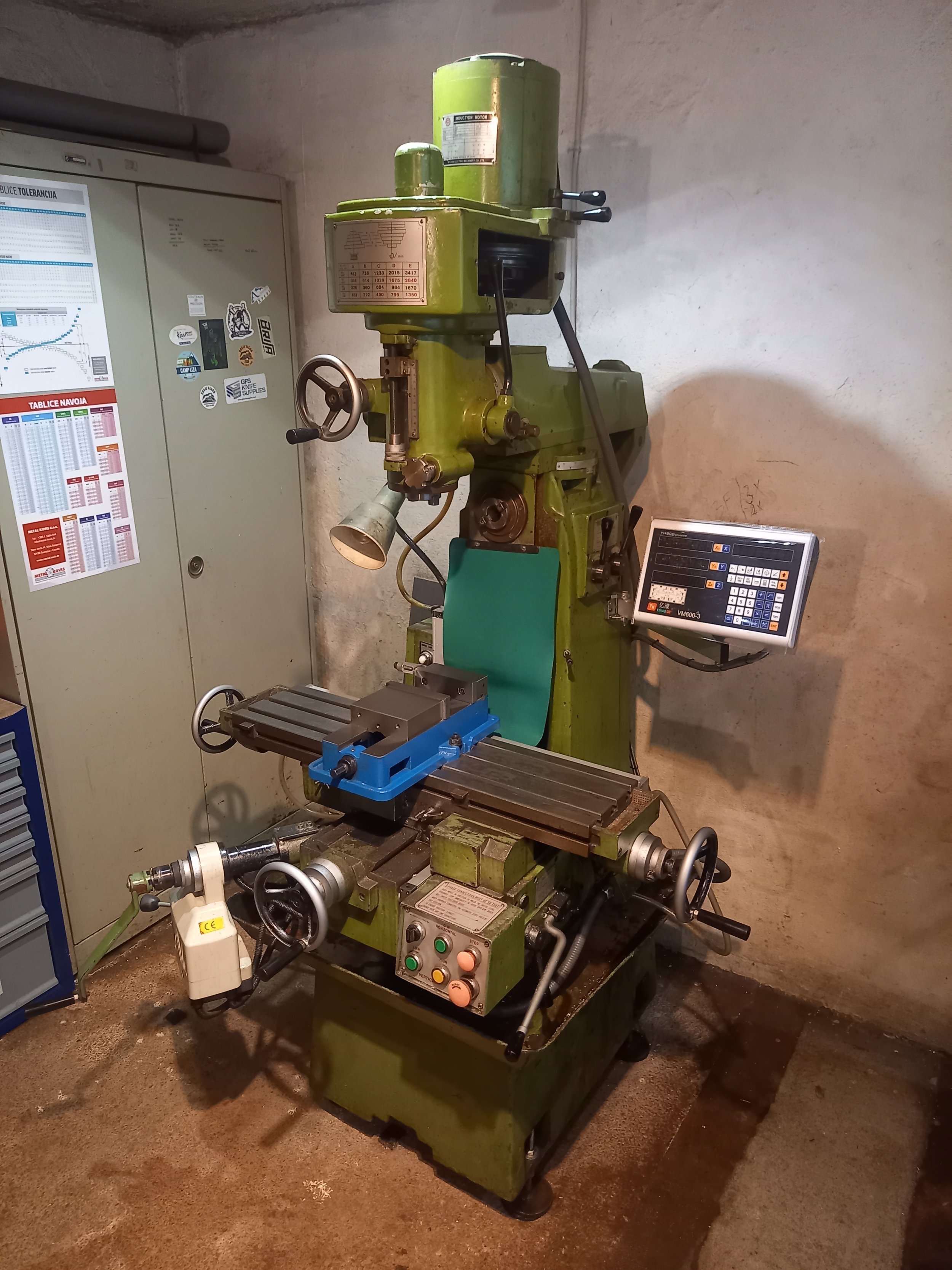 A milling machine in a workshop, with control panel, handwheels, and metal components. The machine is green, seated on a metal base, with a worktable in the front and various controls and assembly parts.