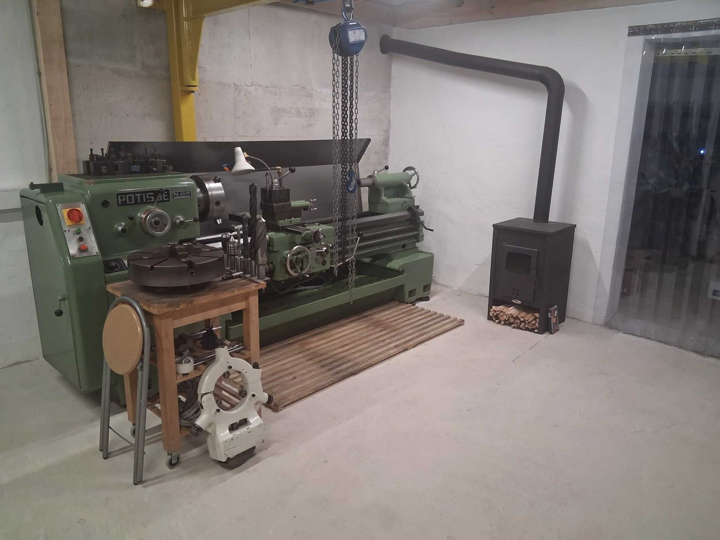 A metalworking workshop with an industrial lathe by Potisje, jib crane hanging from the ceiling, a wood stove. Some wood logs are stacked near the stove.