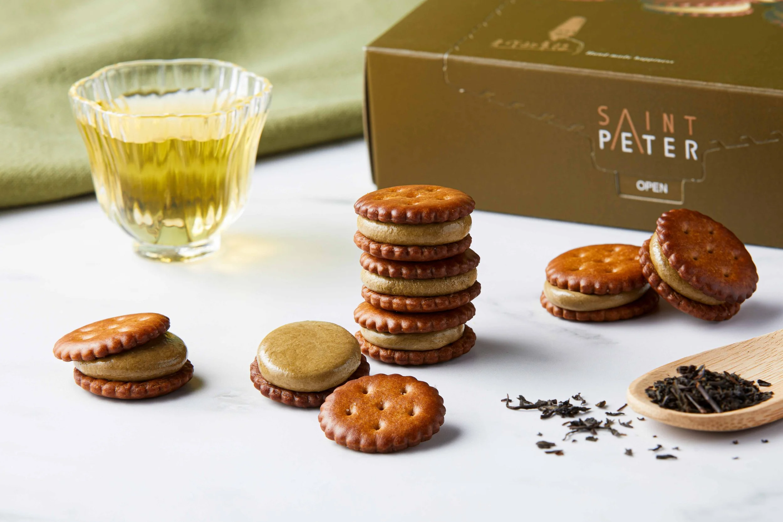 Stacked round cookies with chocolate and vanilla layers next to a glass of yellow beverage and a box labeled Saint Peter on a white surface.