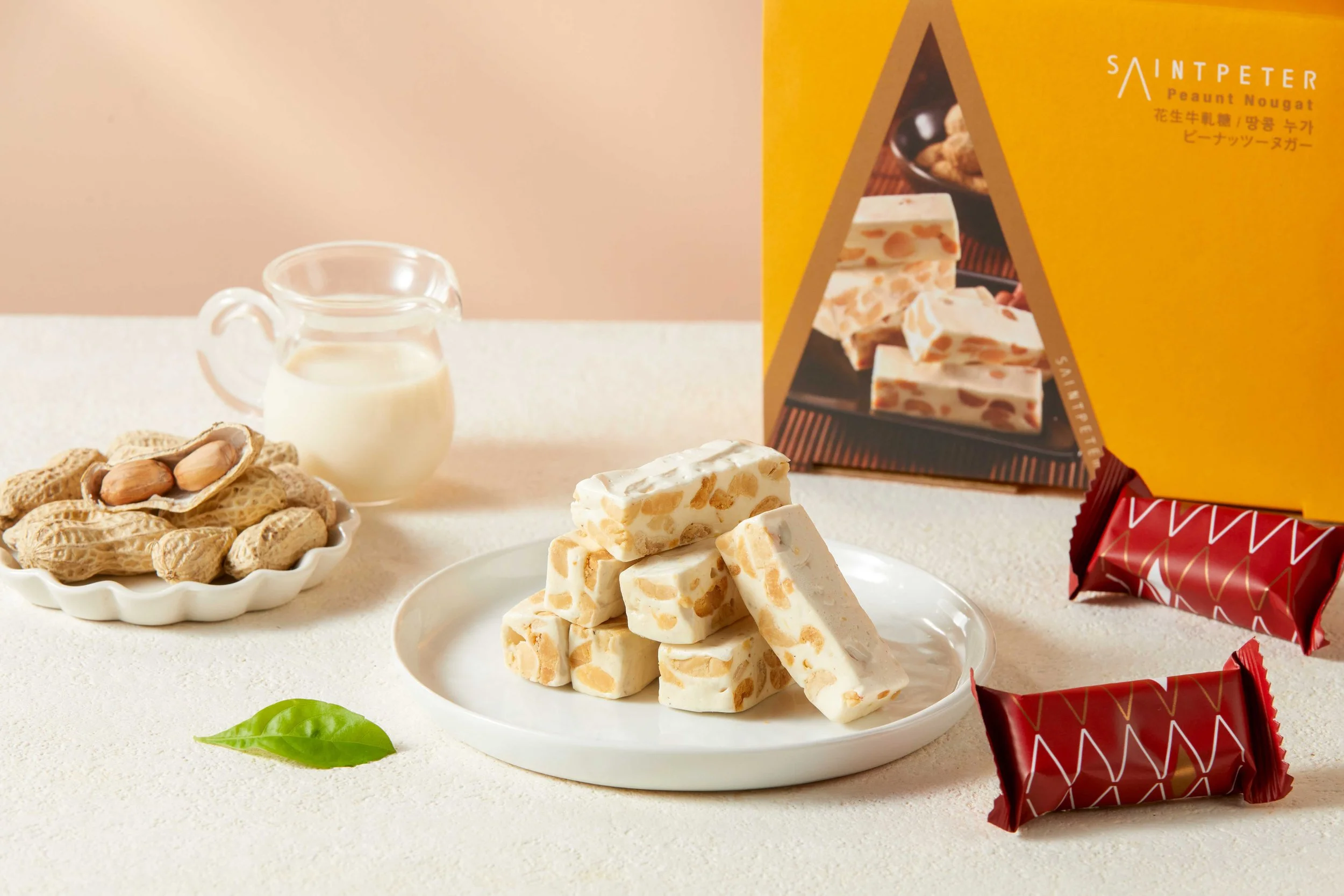 A white plate with caramel peanut nougat pieces, a small bowl of peanuts, a small glass pitcher of milk, a green leaf, and boxes of assorted nougat candies in colorful packaging.