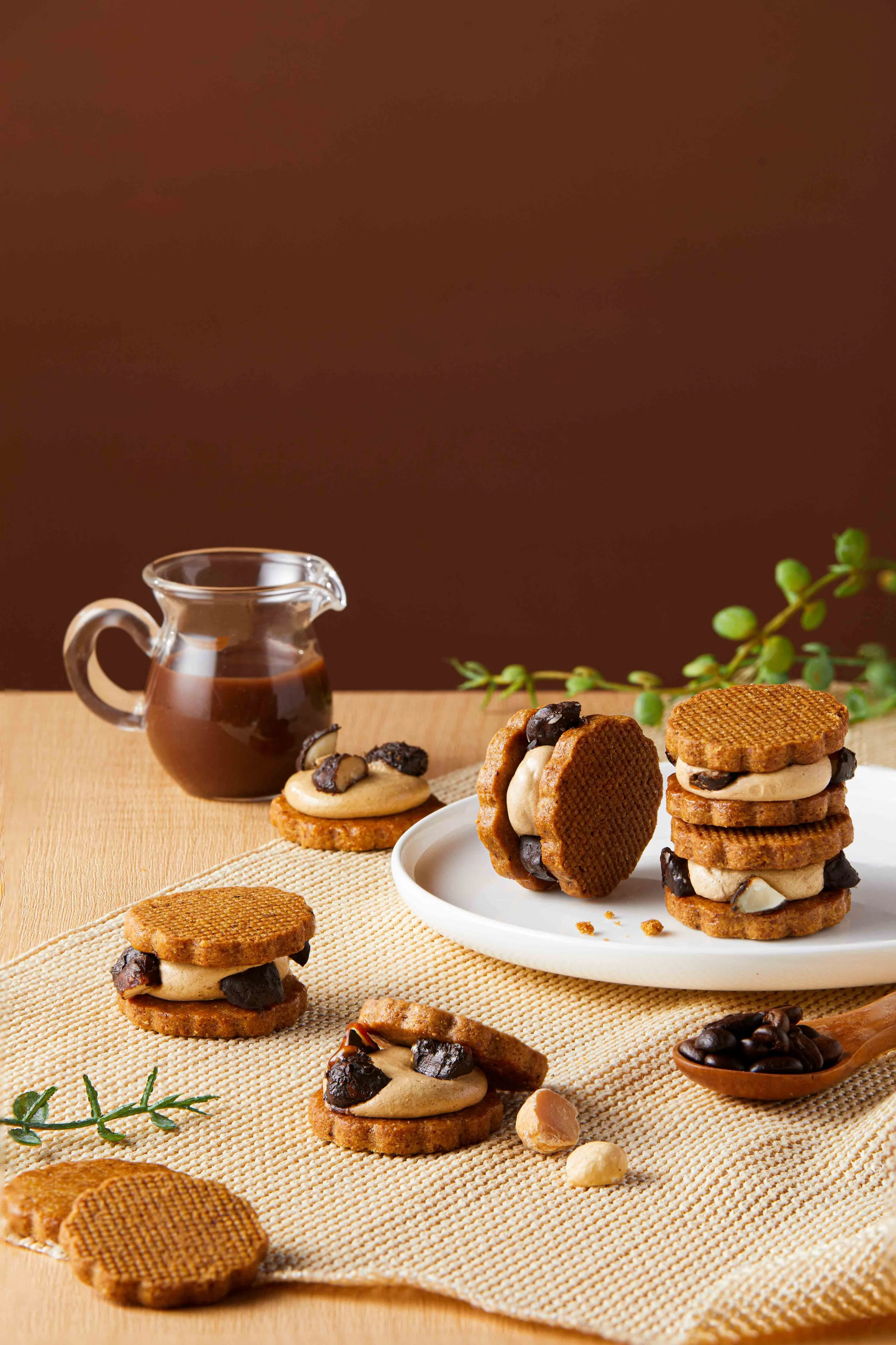 Chocolate sandwich cookies with peanut butter filling and chocolate chips, arranged on a white plate and on a beige woven placemat, next to a glass jar of chocolate sauce, with scattered nuts and coffee beans, and green herbs in the background.