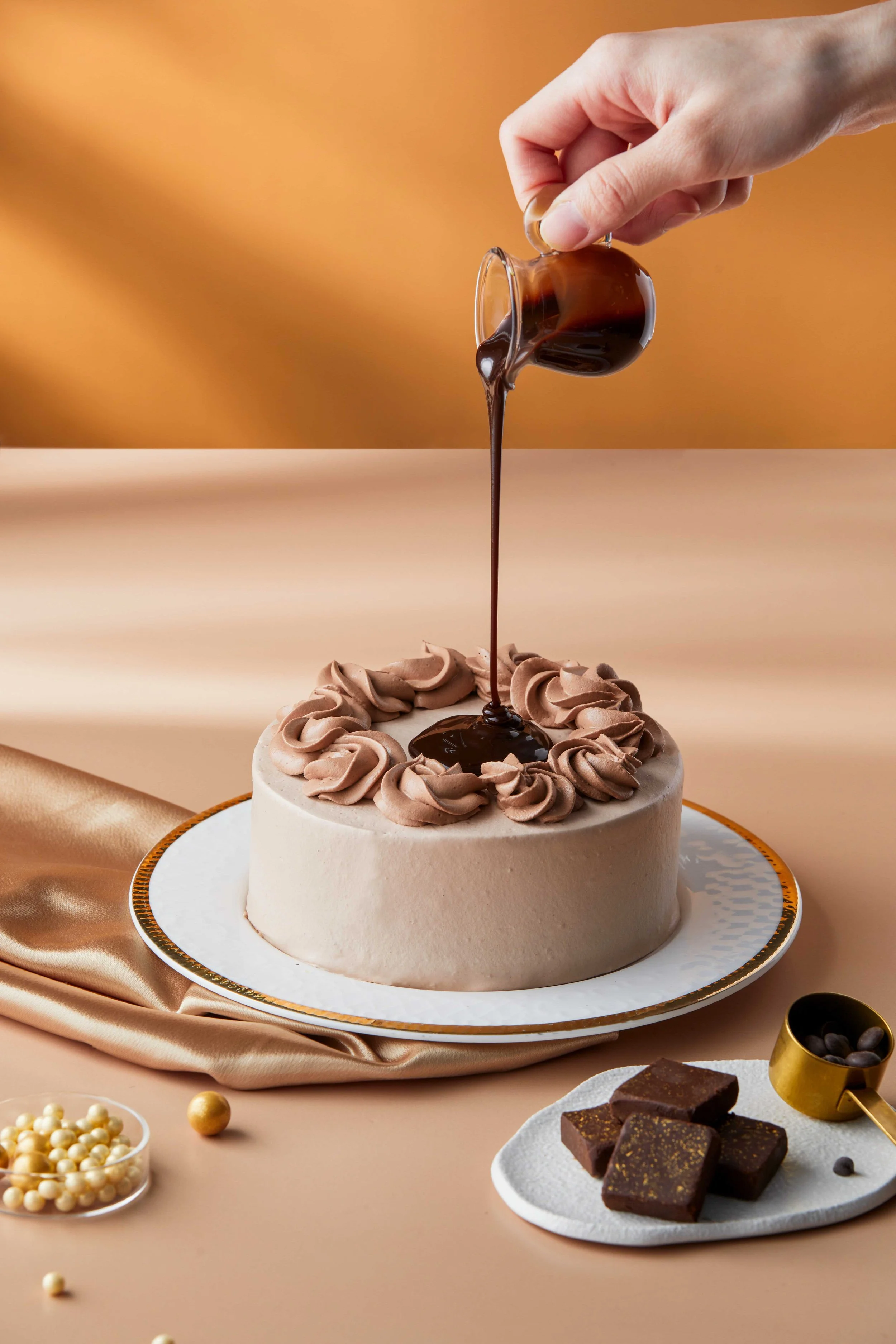 A cake decorated with whipped frosting and chocolate drizzle, with a hand pouring more chocolate onto it.