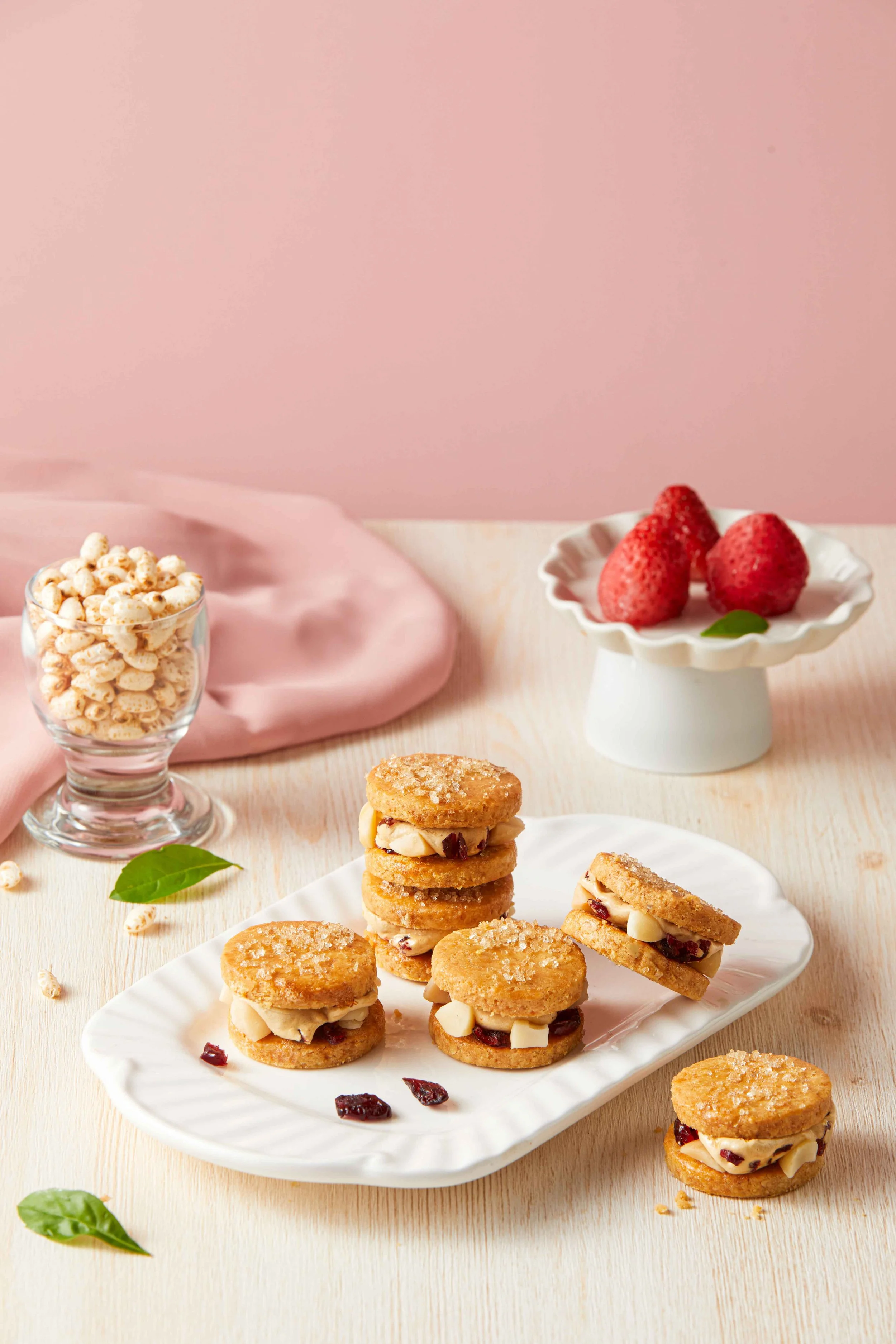 A white plate with small round cookies filled with cream and dried cranberries, some cookies are stacked, on a light wooden table with scattered green leaves and a small jar of popcorn, pink background.