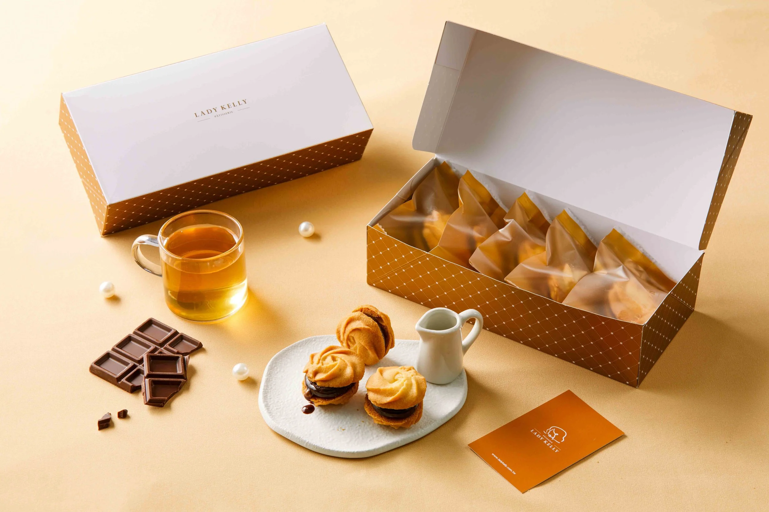 Open box of Lady Kelly cookies on a beige surface with a glass of tea, chocolate pieces, cream, and cookies on a white plate, decorated with pearls and a chocolate bar.