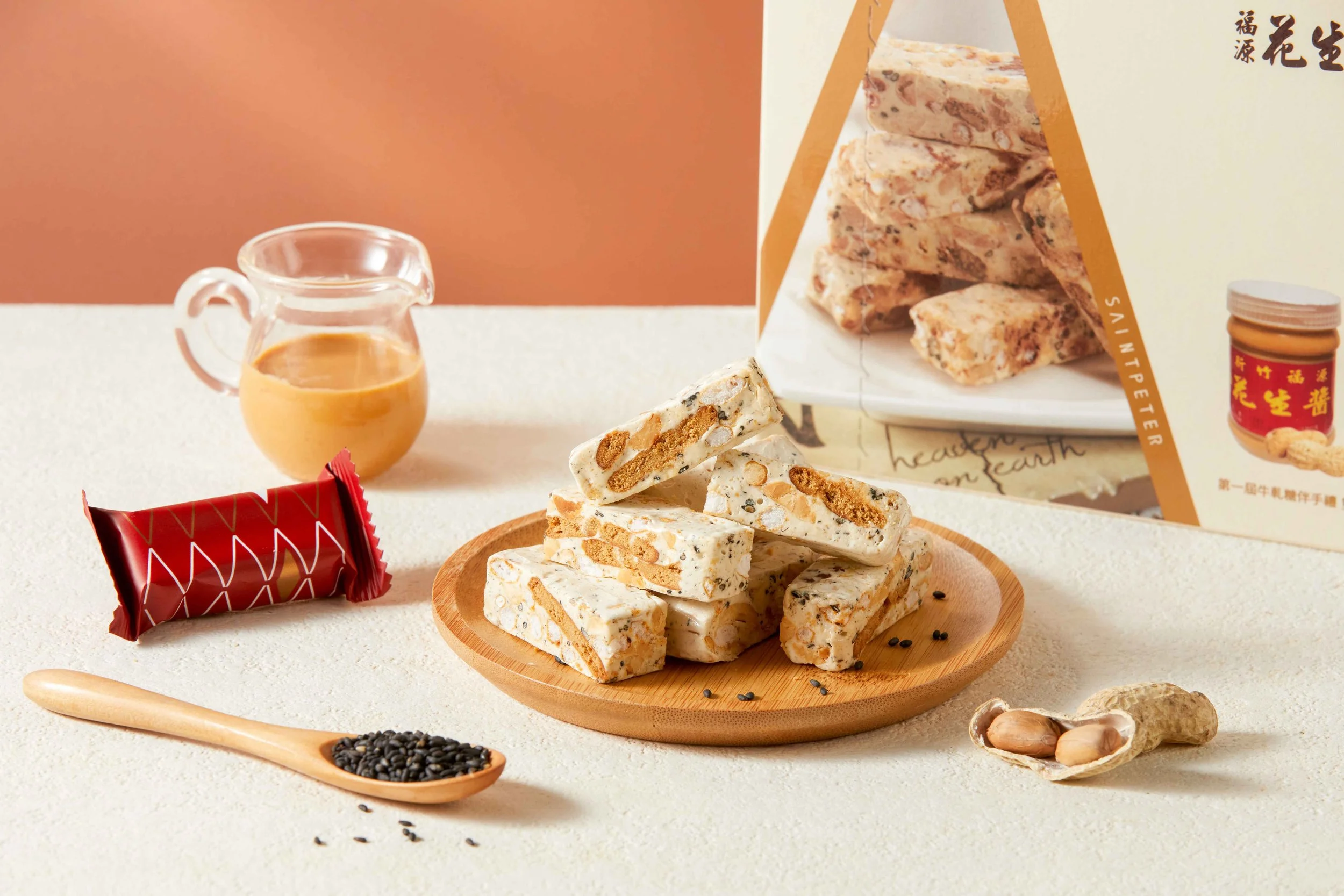 Tray of sliced sesame brittle on a wooden plate, next to a jar of peanut butter, a spoonful of black sesame seeds, and a small hole in the wall of peanut shells.
