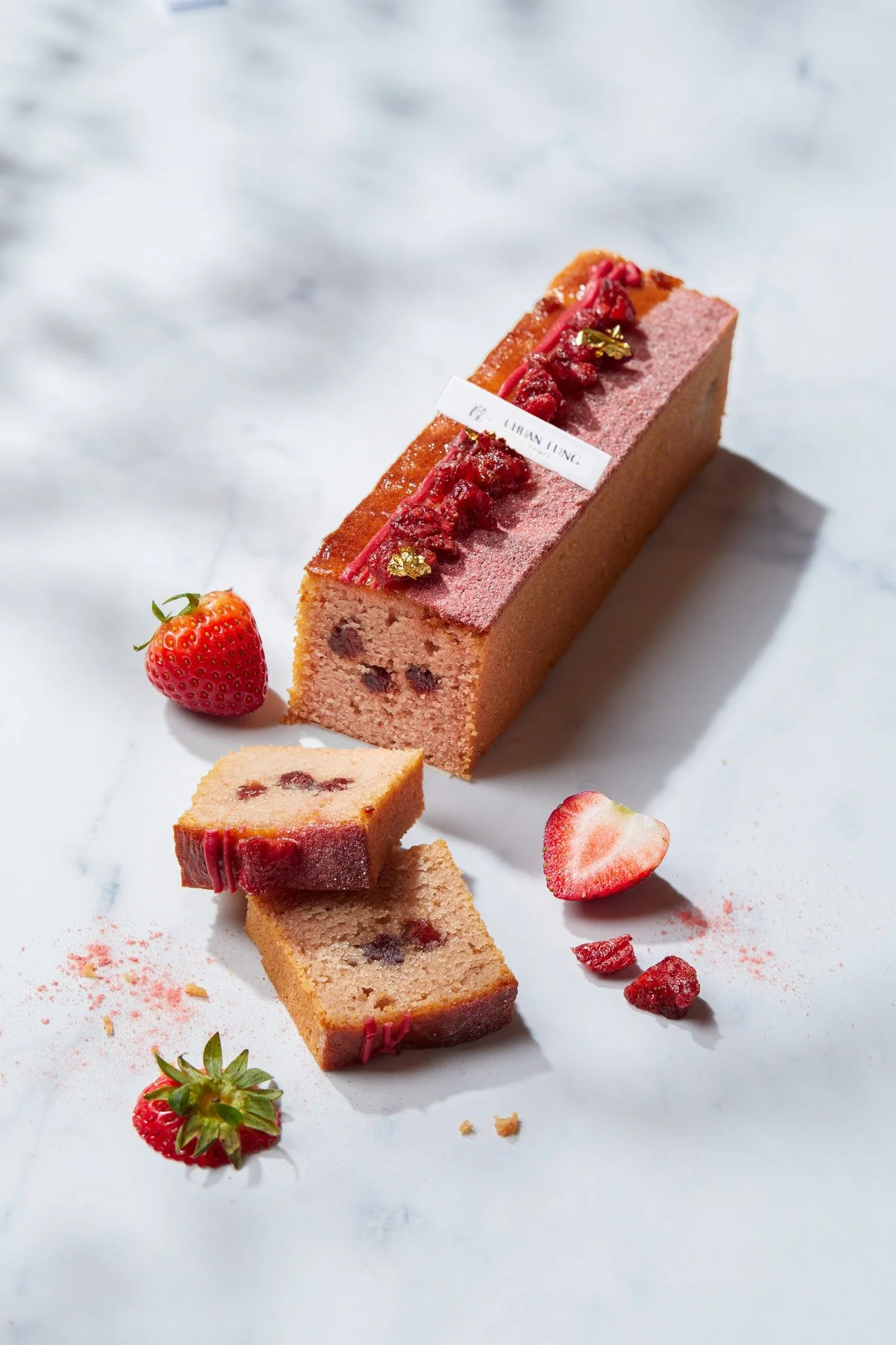 A pink strawberry cake with strawberries and gold leaf garnish, sliced to show its interior with visible strawberry pieces and filling, on a white marble surface with scattered strawberries and crumbs.