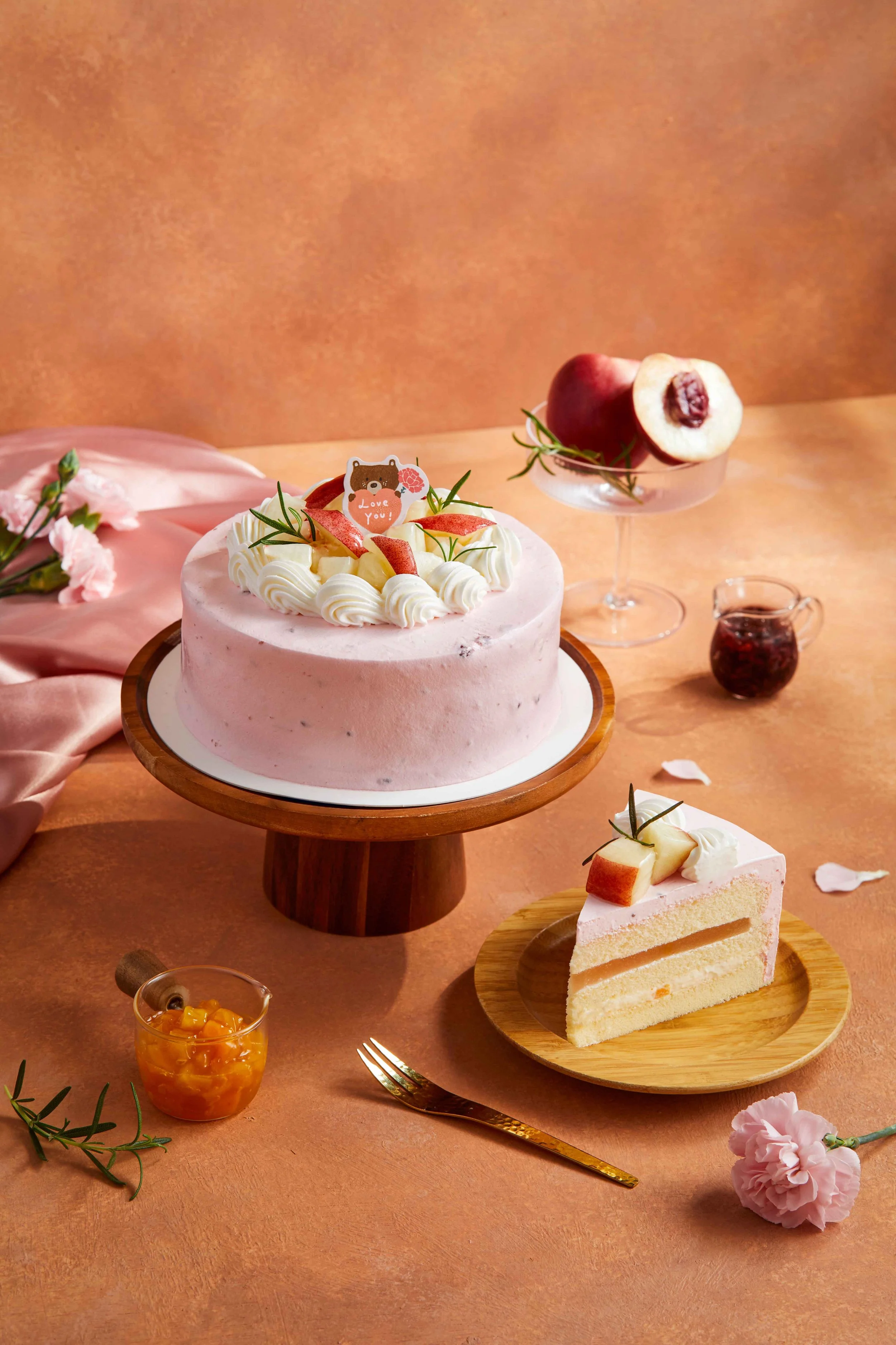 A pink frosted cake decorated with whipped cream, peach slices, and a small bear with a pink flower with the words "Love You". Next to the cake is a slice of layered cake with peach topping on a wooden plate. There is a glass of orange jelly, a small