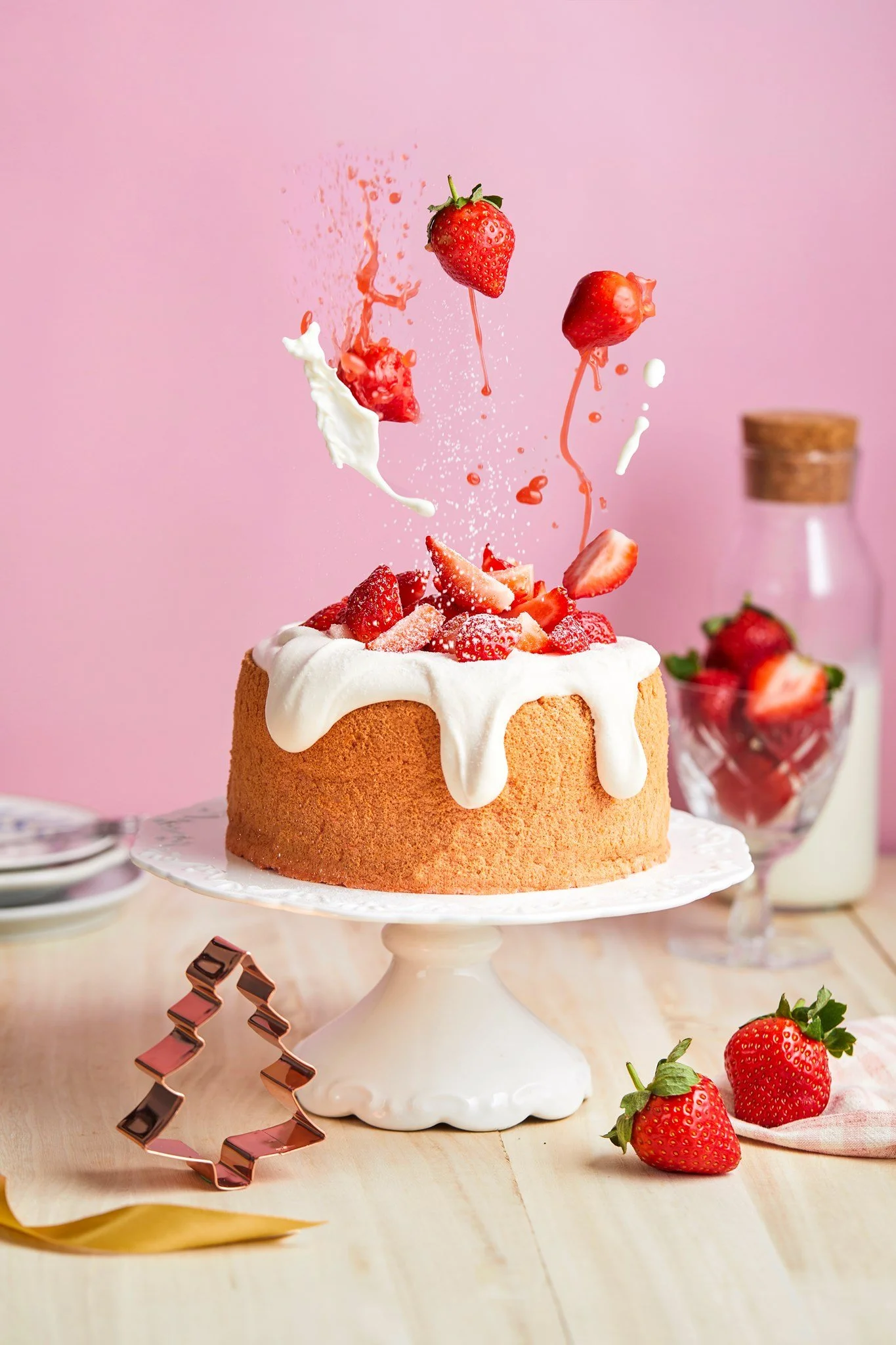 Strawberry cake with whipped cream on a white cake stand, strawberries and chocolate decorations on a wooden table with a pink background.
