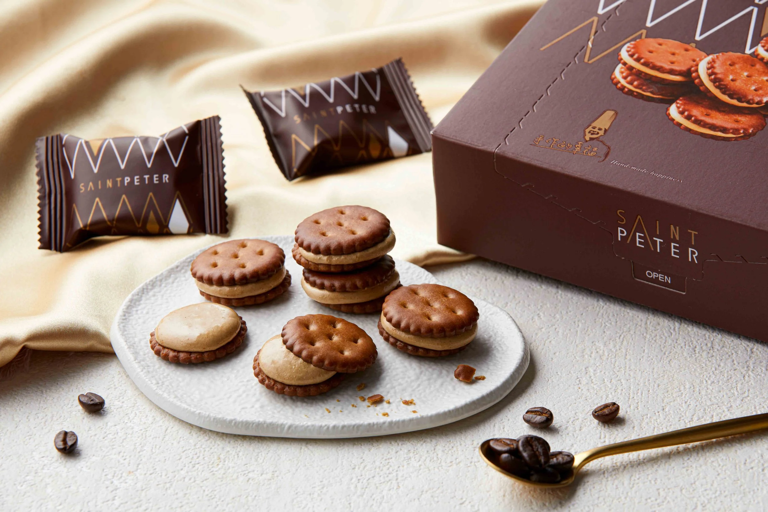 A white plate of round sandwich cookies with cream filling, scattered coffee beans, a chocolate box with cookies on it, and two wrapped packages labeled Saint Peter, on a cream satin fabric.