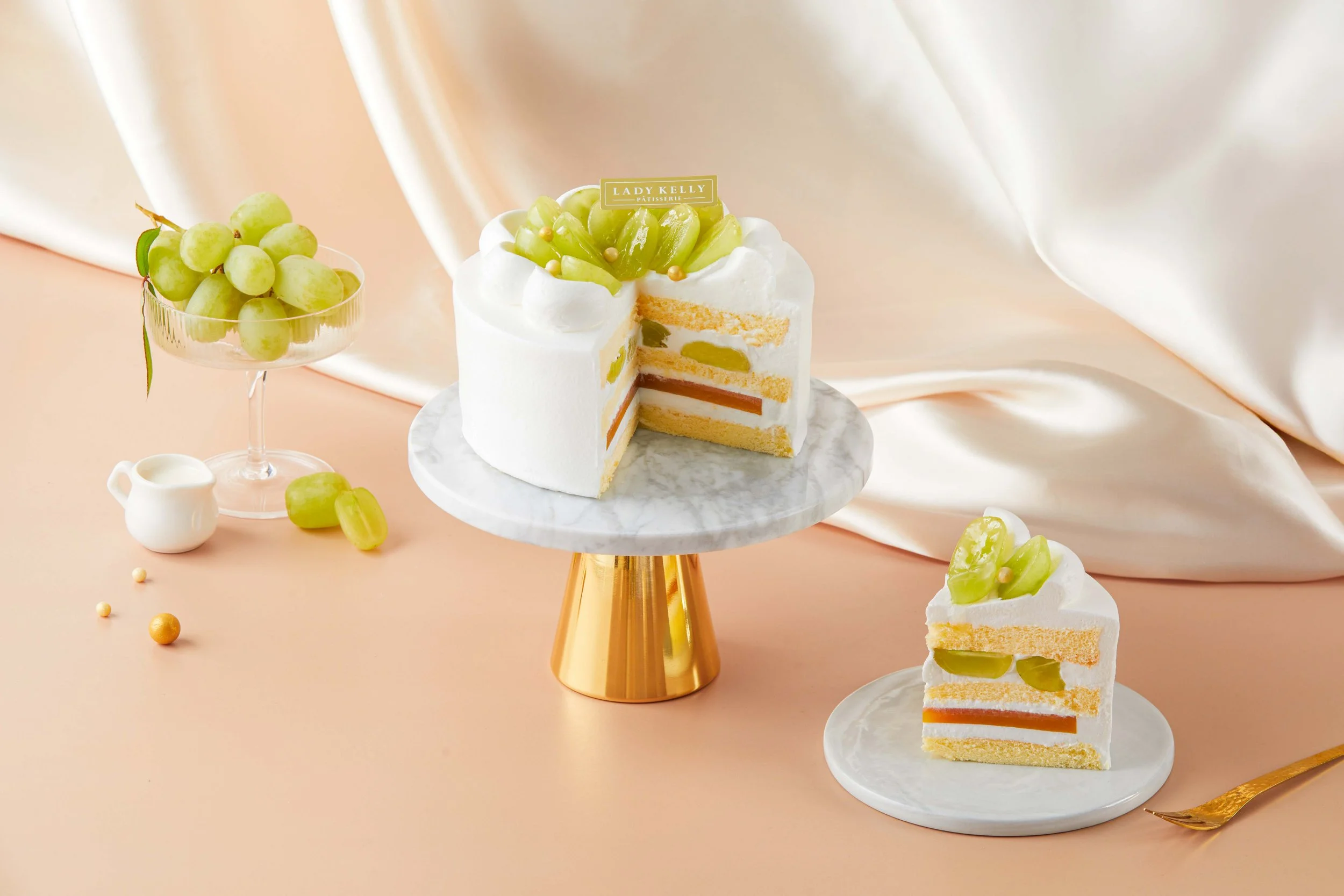 A white cake with green grapes on top, layered with cream and fruit filling, displayed on a marble cake stand, with additional slices on white plates. To the left, a glass bowl of green grapes, a small white pitcher, and scattered grapes and small ye
