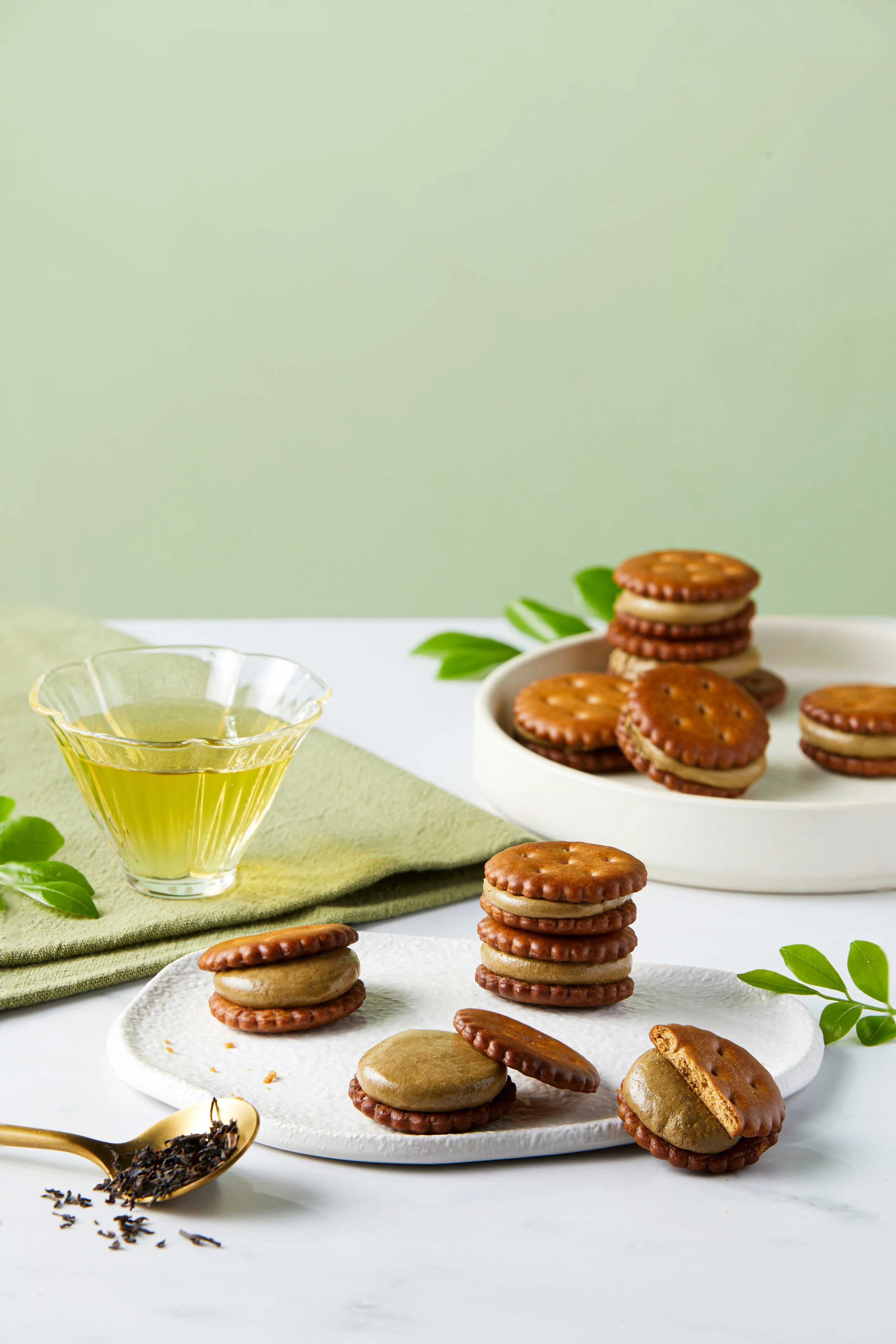Assorted sandwich cookies with cream filling arranged on white plates, a glass bowl of tea, and fresh green leaves on a light green cloth.
