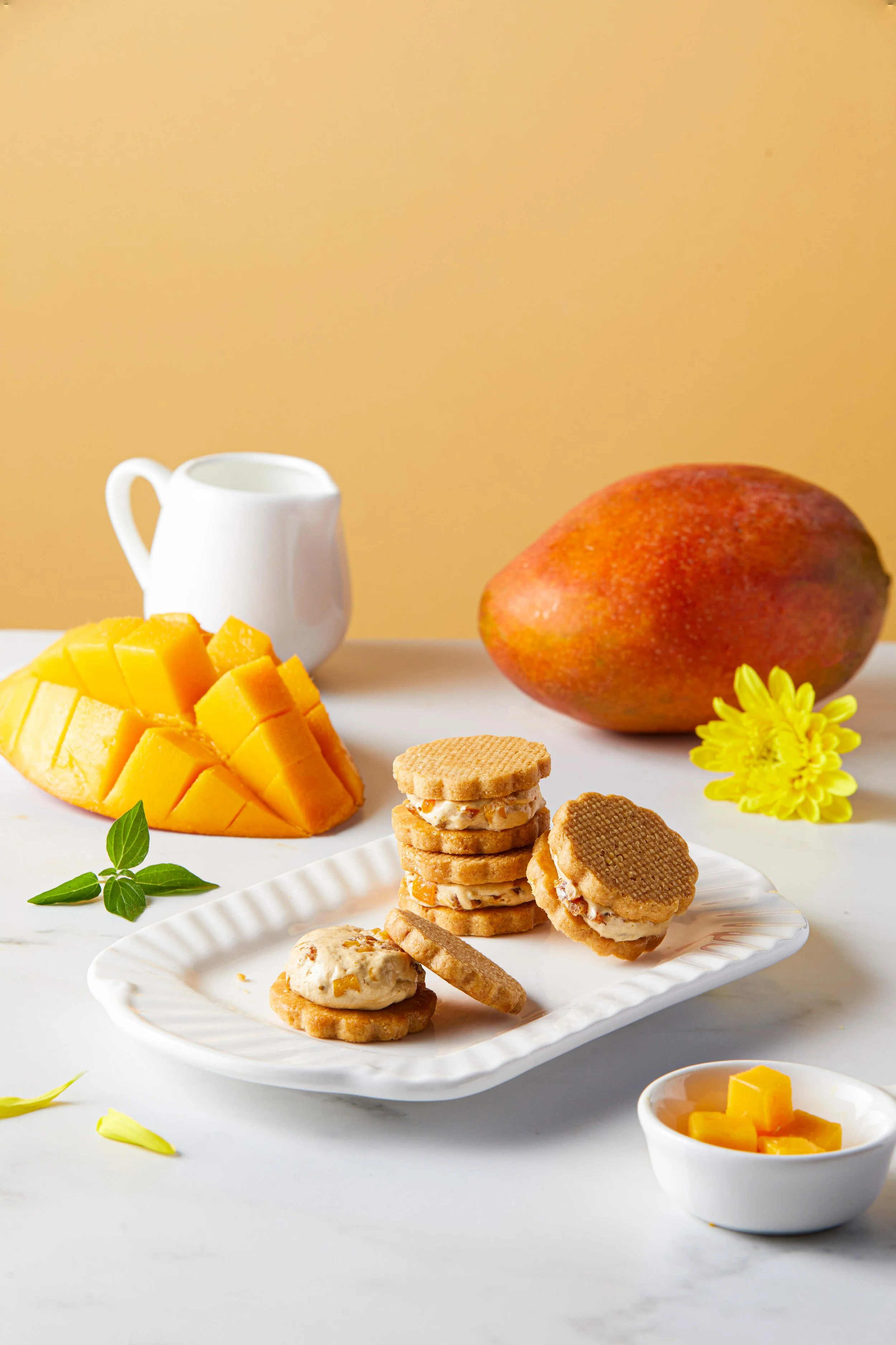 Close-up of a plate of cookies with a stack of sandwich cookies and a single cookie topped with fruit preserves, garnished with mint leaves, on a white surface with a background of a whole mango, a white milk pitcher, a small bowl of mango pieces, an