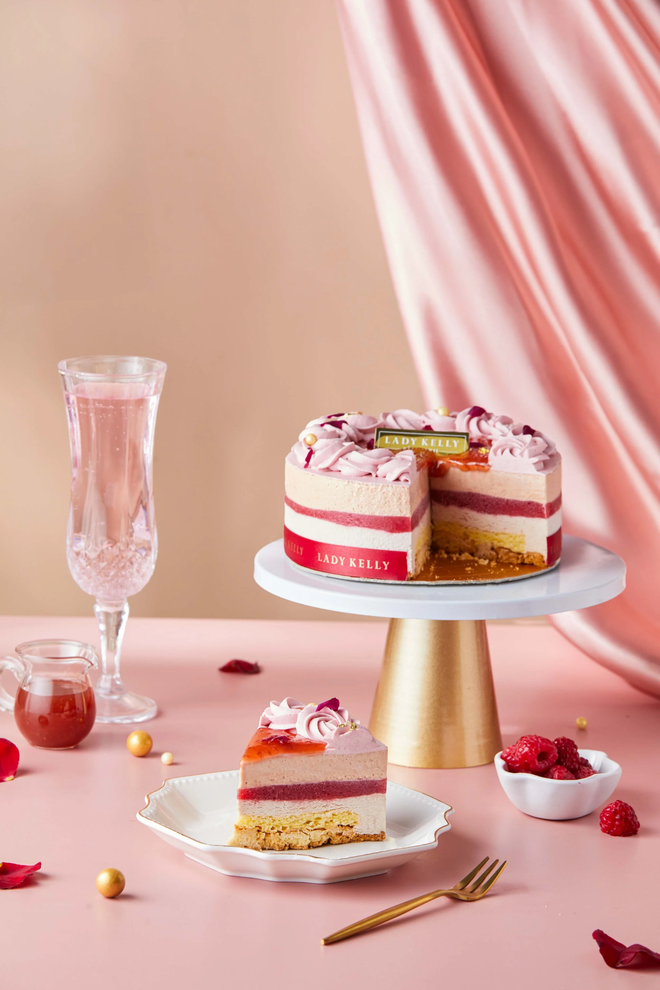 A pink cake with pink and white icing, topped with gold pearls and a "Lady Kelly" label, is on a white cake stand. A slice is cut and placed on a white plate. Pink and gold decorations, fresh raspberries, and a glass of pink drink are arranged on a p