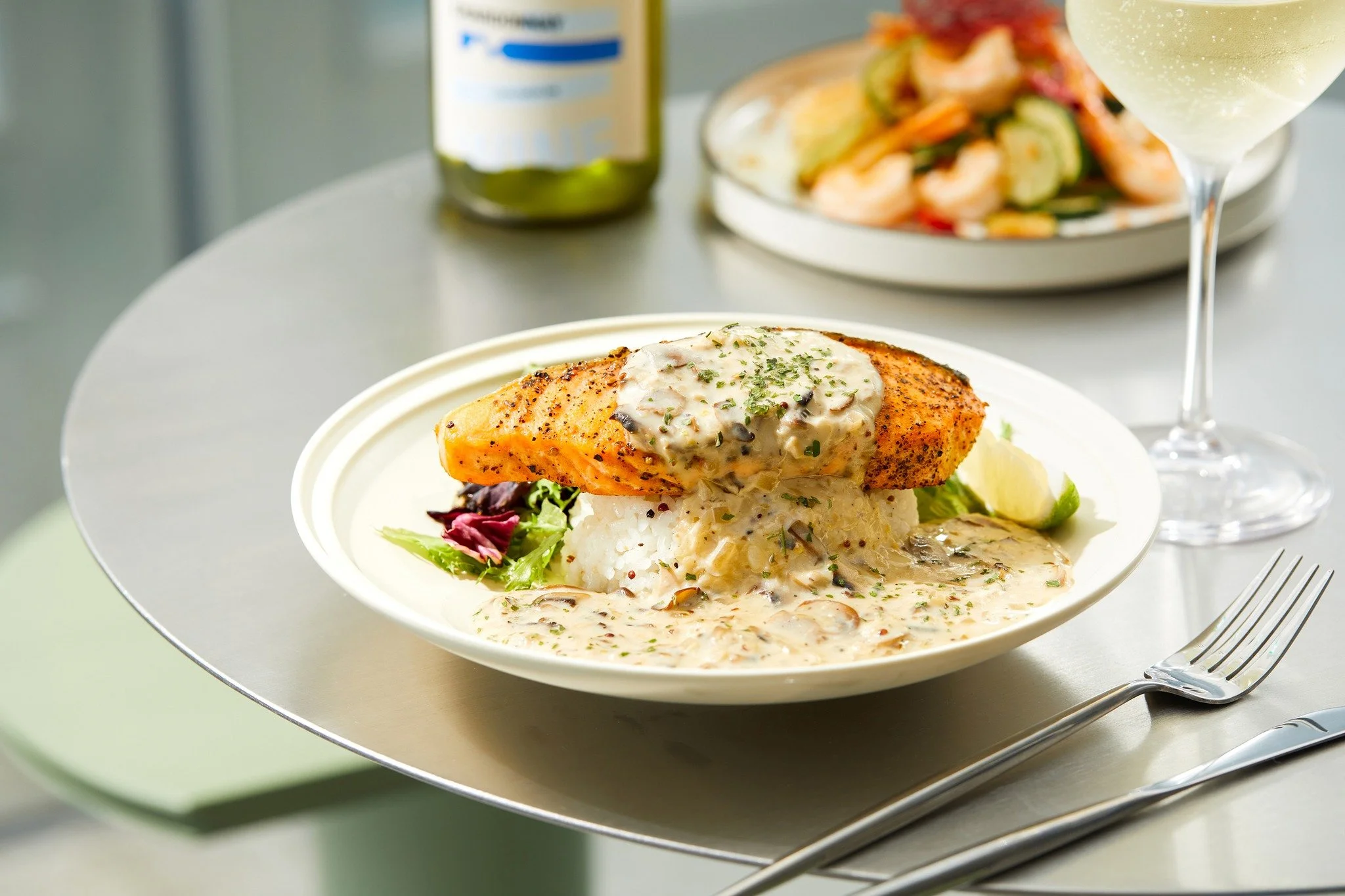 A plated dish featuring a cooked fillet of salmon topped with a creamy mushroom sauce, served with rice and garnished with fresh herbs, along with a wedge of lemon, on a white plate. In the background, there's a glass of white wine, a plate of mixed salad, and a bottle of green liquid, possibly olive oil or dressing, all on a metal serving tray.