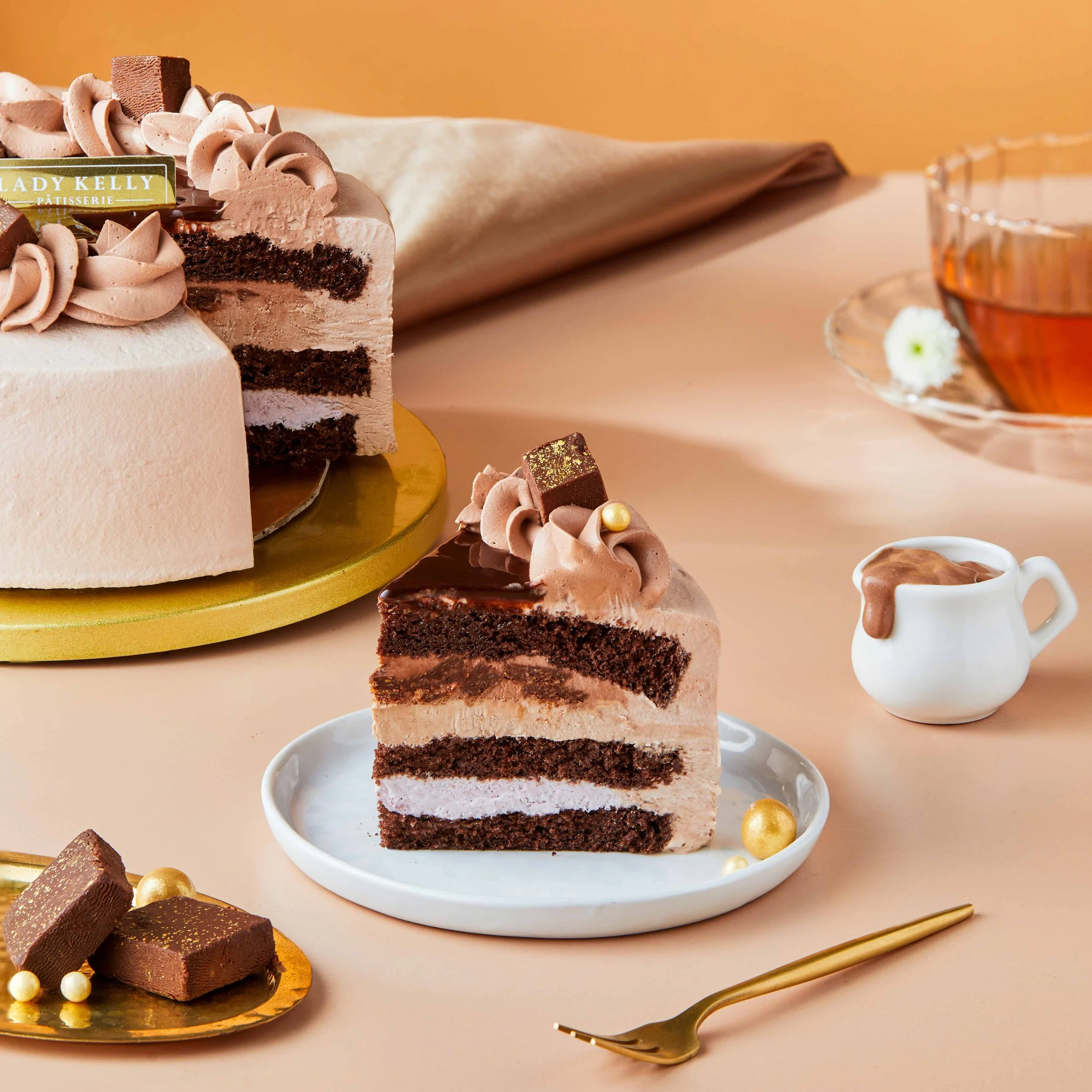 A slice of layered chocolate and pink cake with chocolate frosting and gold pearl decorations on a white plate, with chocolate desserts and gold utensils in the background.