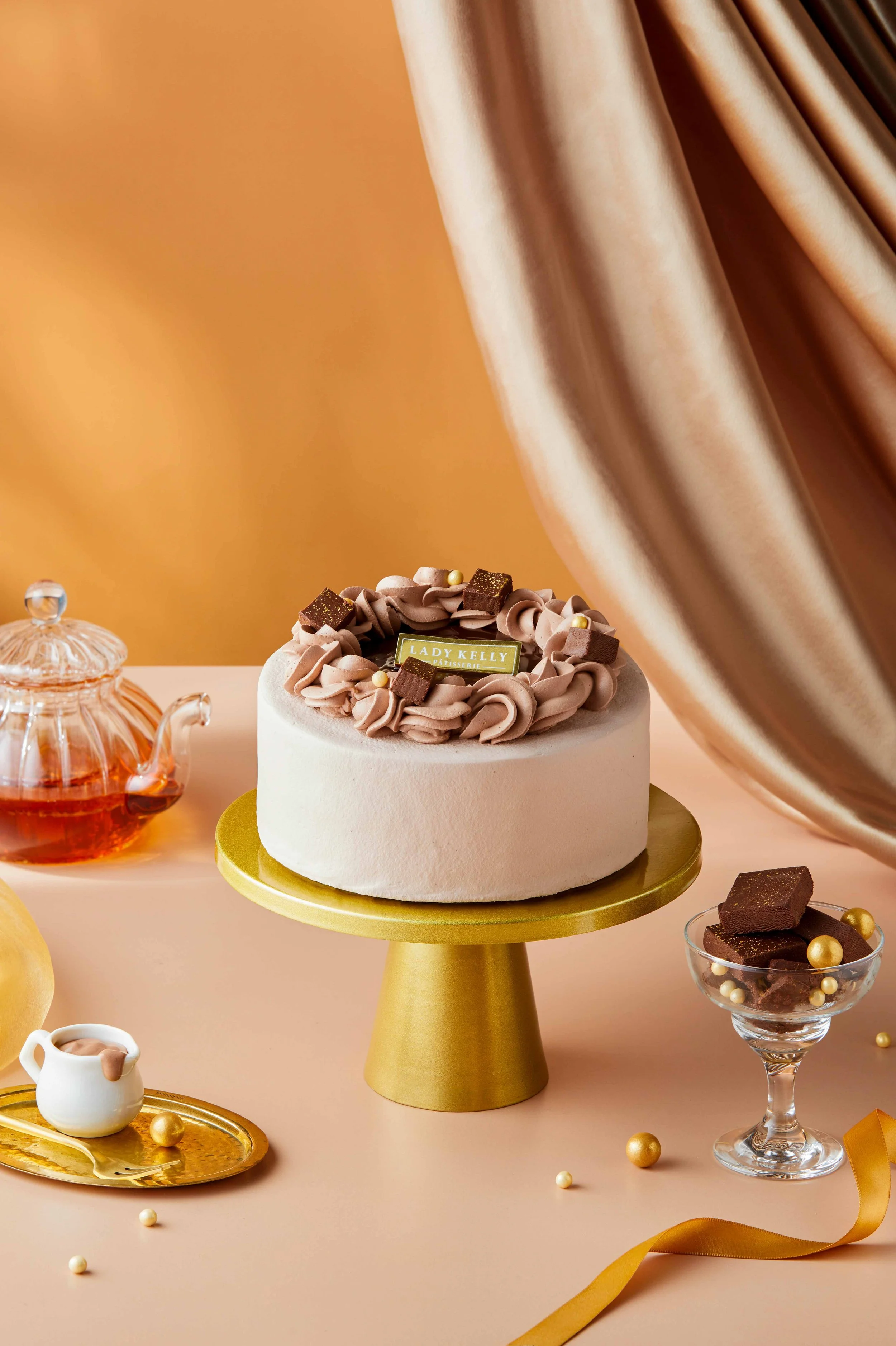 A round frosted cake topped with swirls of brown icing, chocolate pieces, and gold and pearl decorations, displayed on a gold cake stand with a gold label reading 'Lady Kelly'