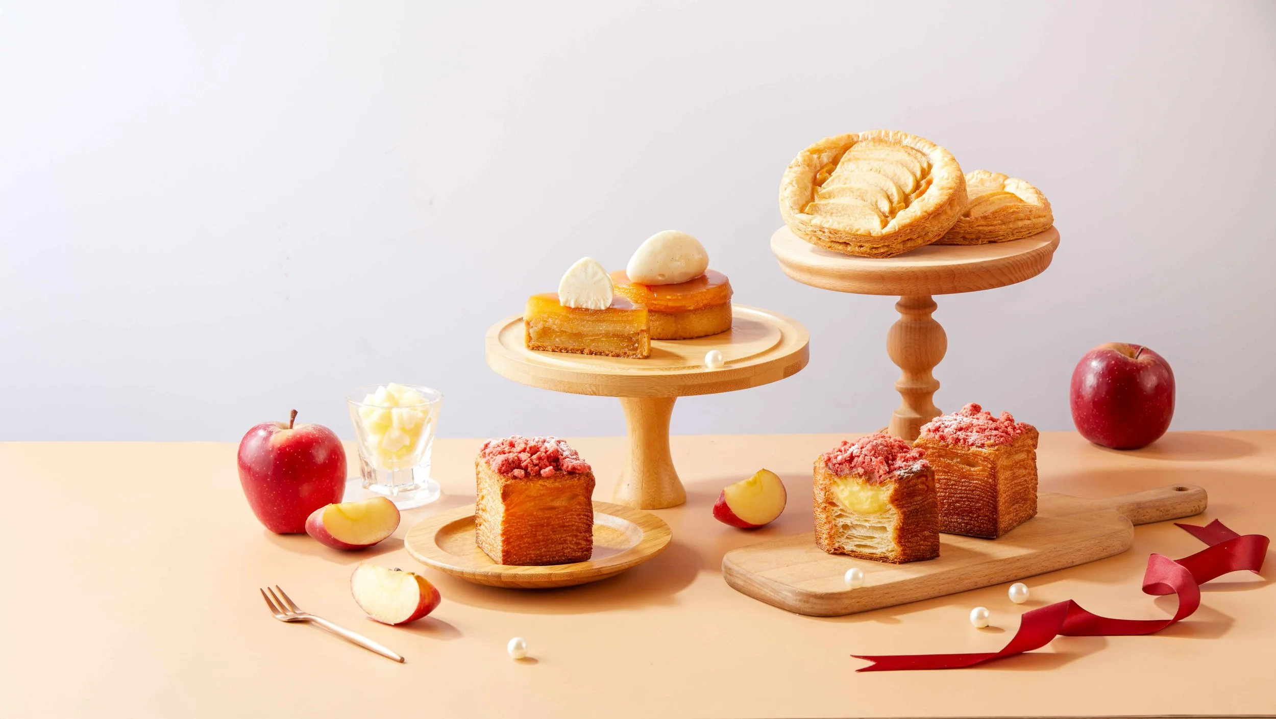 An assortment of baked goods, including apple Tarts and cakes, displayed on wooden stands, with apples, a glass of white chocolate, and decorative elements on a light-colored table against a plain background.