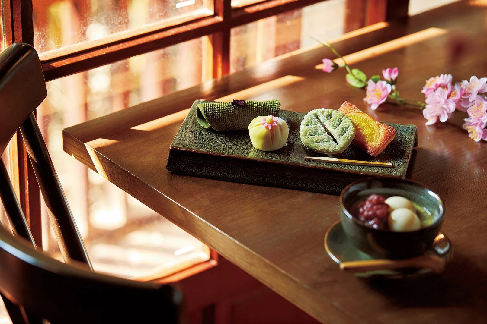 A traditional Japanese tea and dessert set on a wooden table, featuring a black bowl with small round mochi and red bean paste, a rectangular plate with green tea-themed sweets including a matcha cookie, a white bean cake, and a green sweet, all decorated with pink flower accents, with pink flowers lying nearby and sunlight streaming through a wooden window.