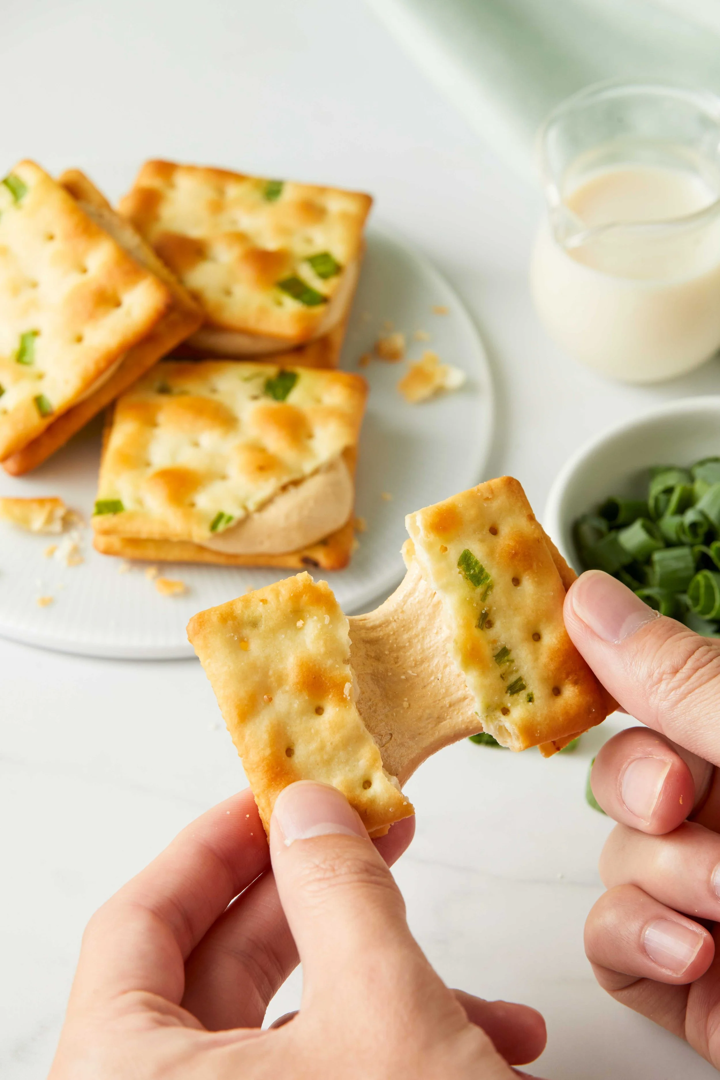 Close-up of hands holding a saltine cracker with green onions, with more crackers, a small bowl of sliced green onions, a glass of milk, and a plate of crackers in the background.