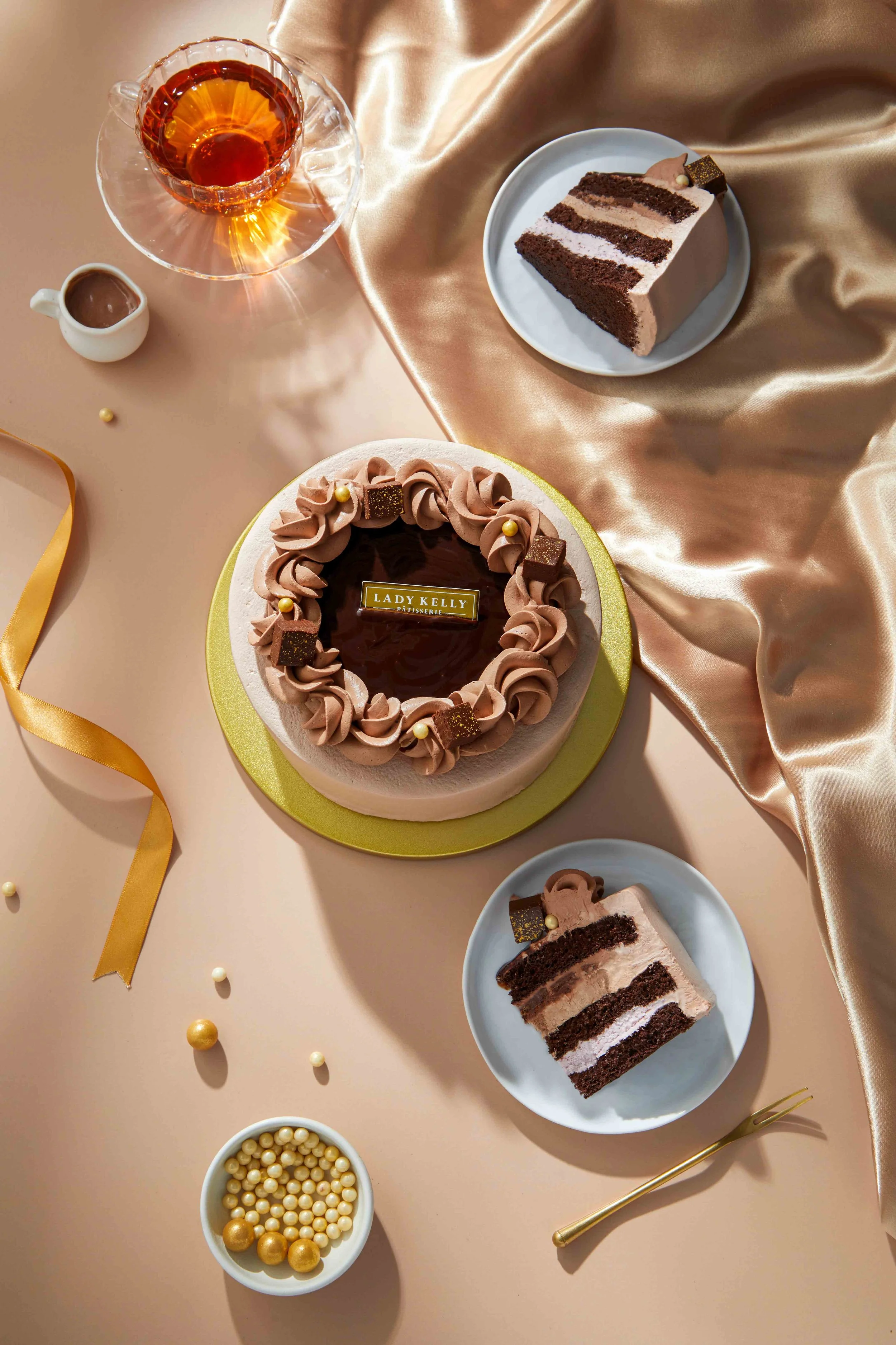 A table with two slices of layered chocolate cake on blue plates, a chocolate birthday cake with pink frosting and gold and chocolate decorations, a glass cup of tea on a glass saucer, small bowls of white and gold spherical candies, and decorative g