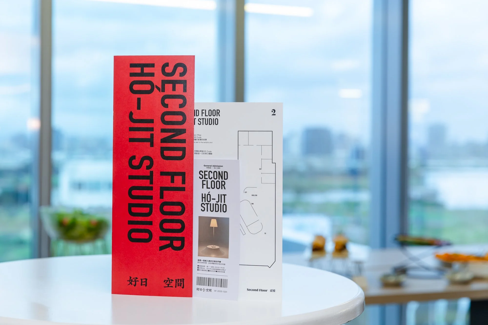 Red sign with black text that reads 'STOP COVID-19 FLY' and a white and black floor plan with some words including 'Second Floor' and and some Chinese characters, stand on a white table with a blurred interior background featuring large windows and cityscape.