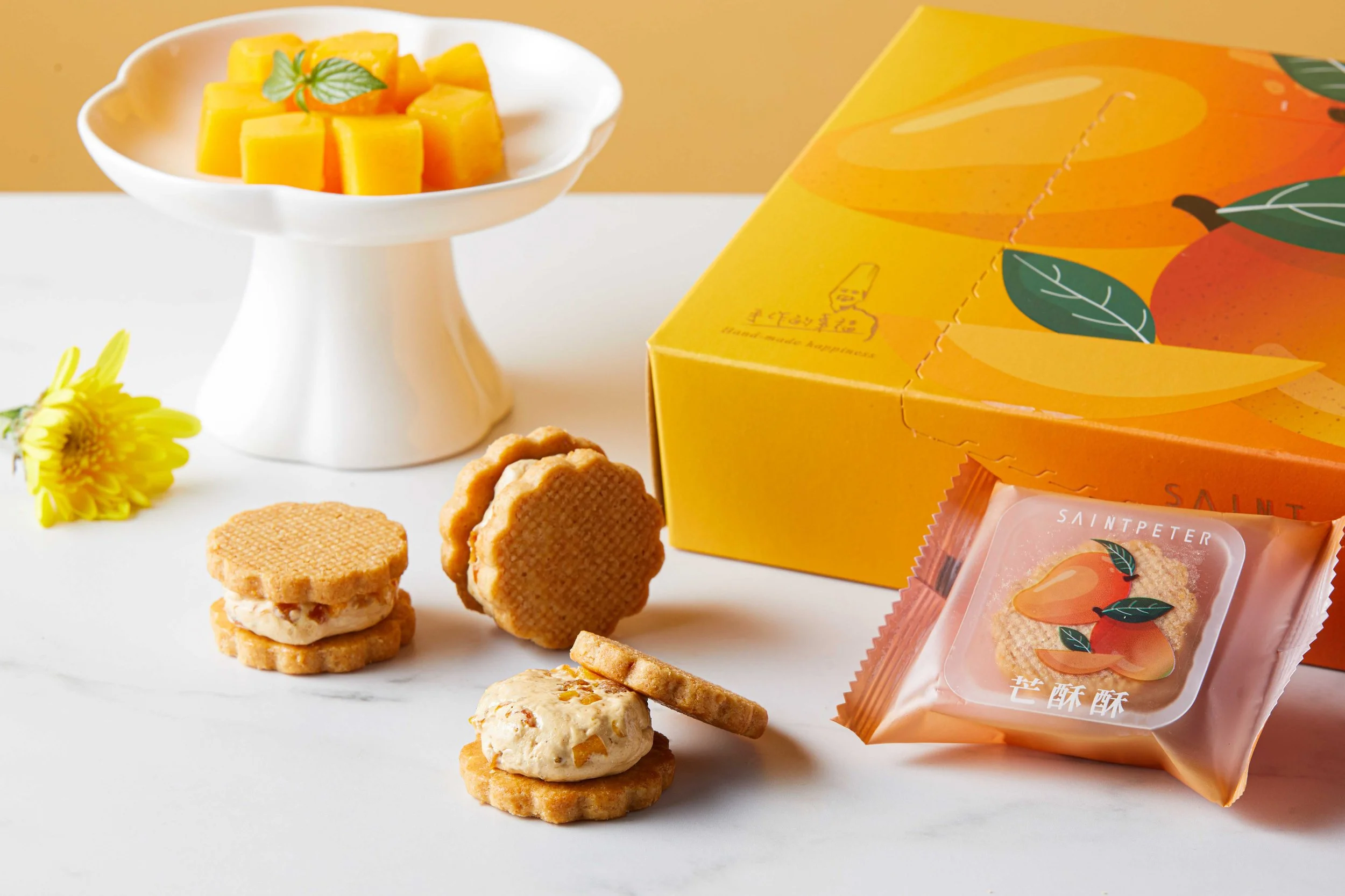 Assorted cookies, a white cake stand with mango chunks garnished with a mint leaf, a yellow box with a mango and green leaves design, and mango-flavored snack packets on a white surface.