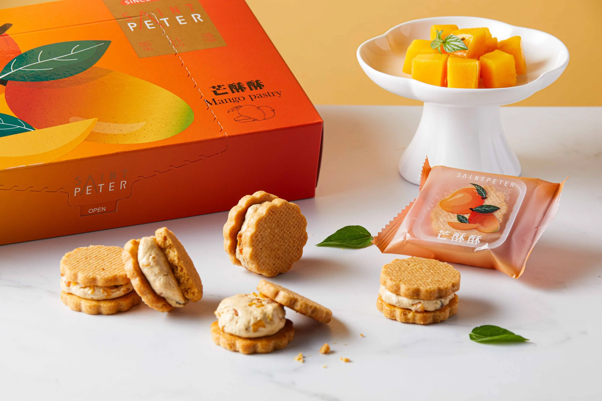 Box of Saint Peter mango pastries with some shortbread cookies and pieces of cookies on a white surface, garnished with green leaves, against a plain background.