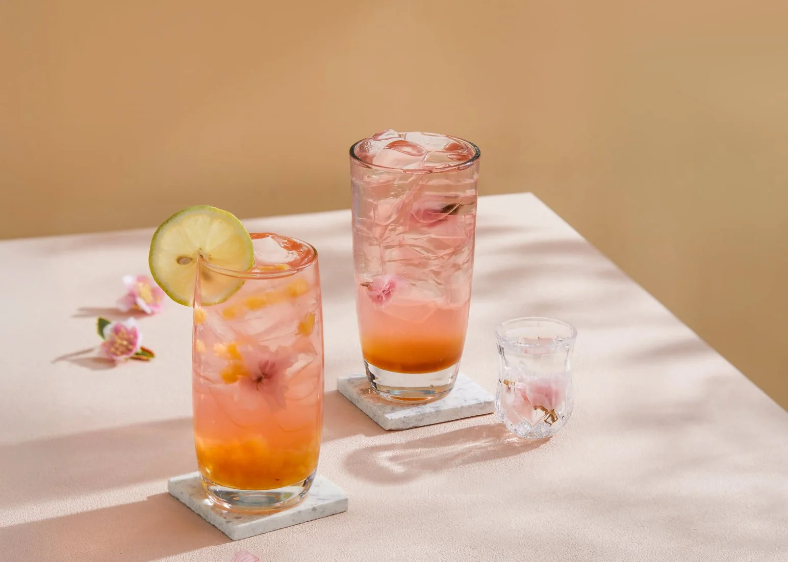 Two pink, floral-infused drinks with ice, one garnished with a lemon slice, placed on marble coasters on a light pink surface, with pink flowers nearby and a small shot glass beside them.