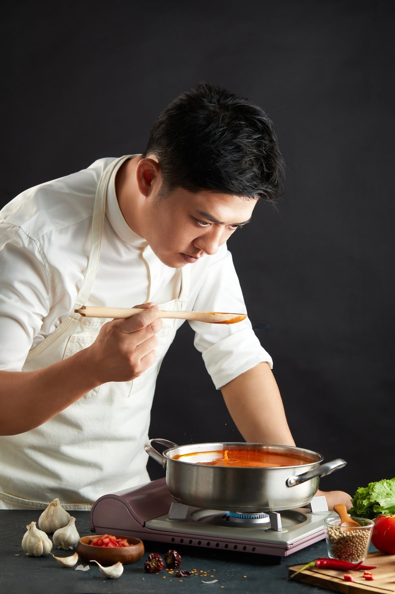 A man cooking on a portable stove, stirring a pot of tomato-based sauce, surrounded by garlic, chopped tomatoes, and various spices.