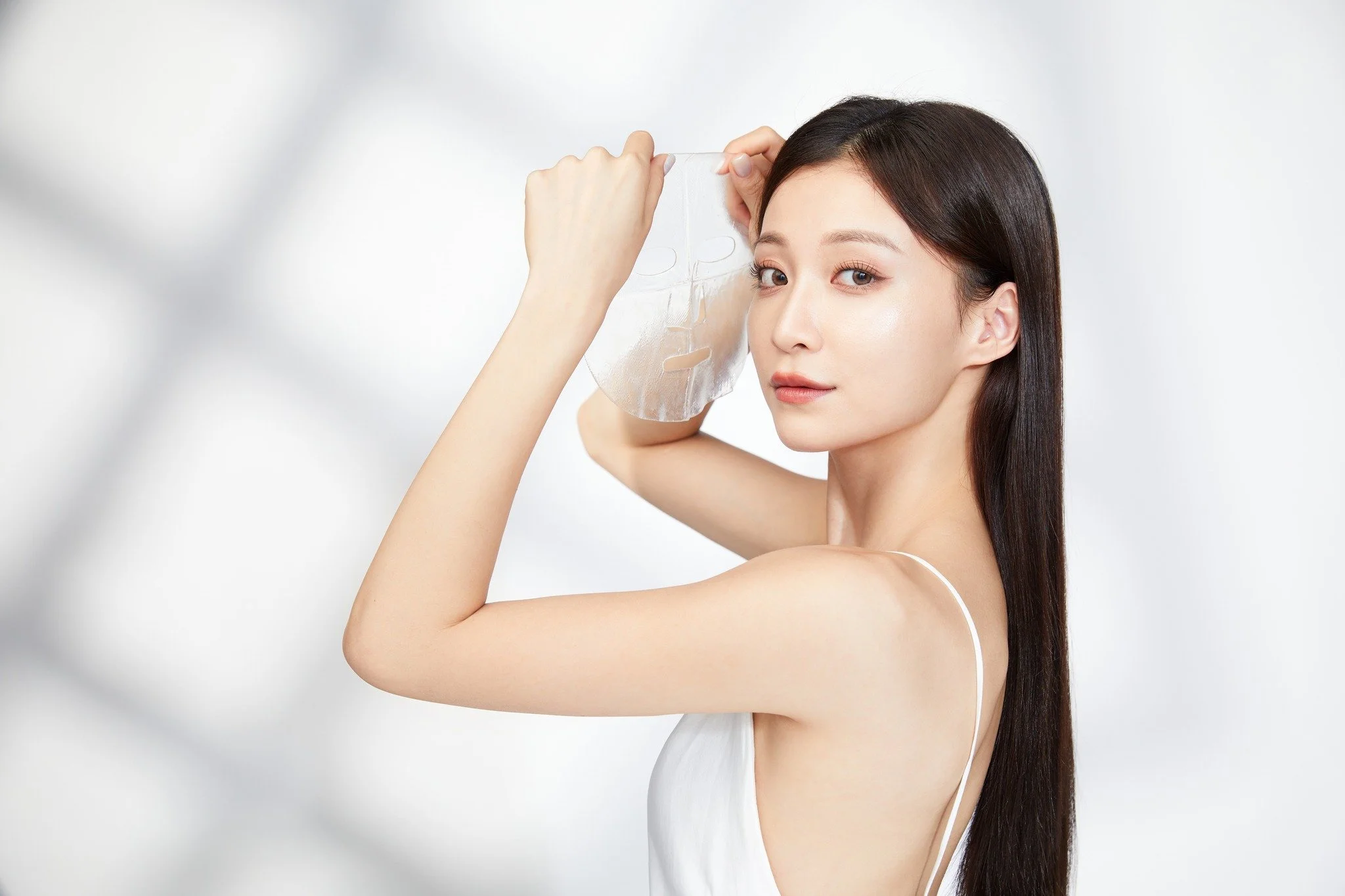 A woman with long dark hair and fair skin applies a facial sheet mask, looking at the camera against a plain light background.