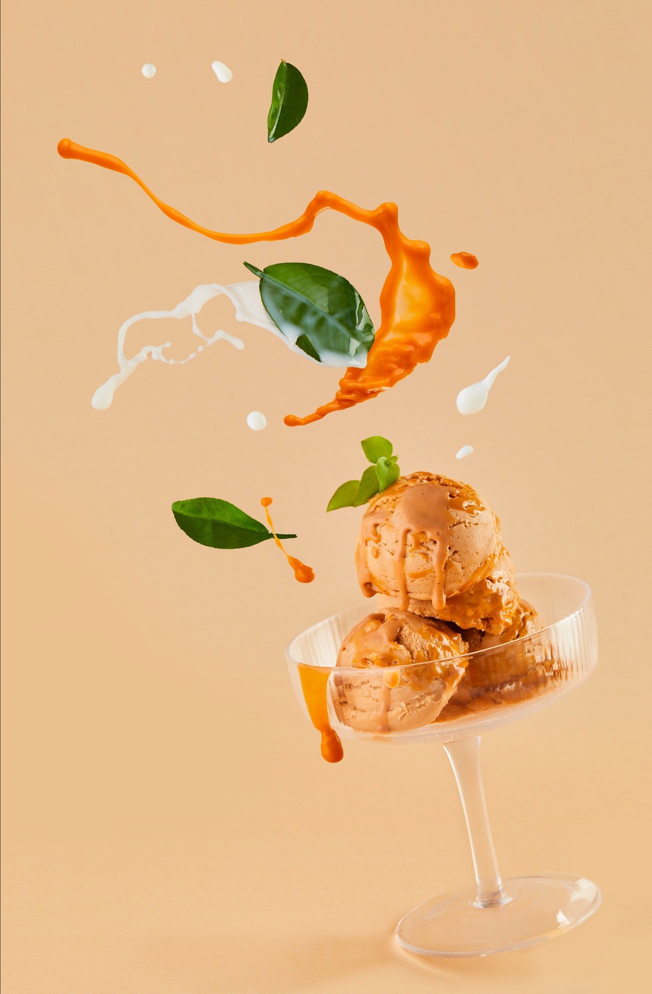 Scoops of caramel ice cream topped with caramel syrup in a glass dessert cup, with splash and green leaves in the background.