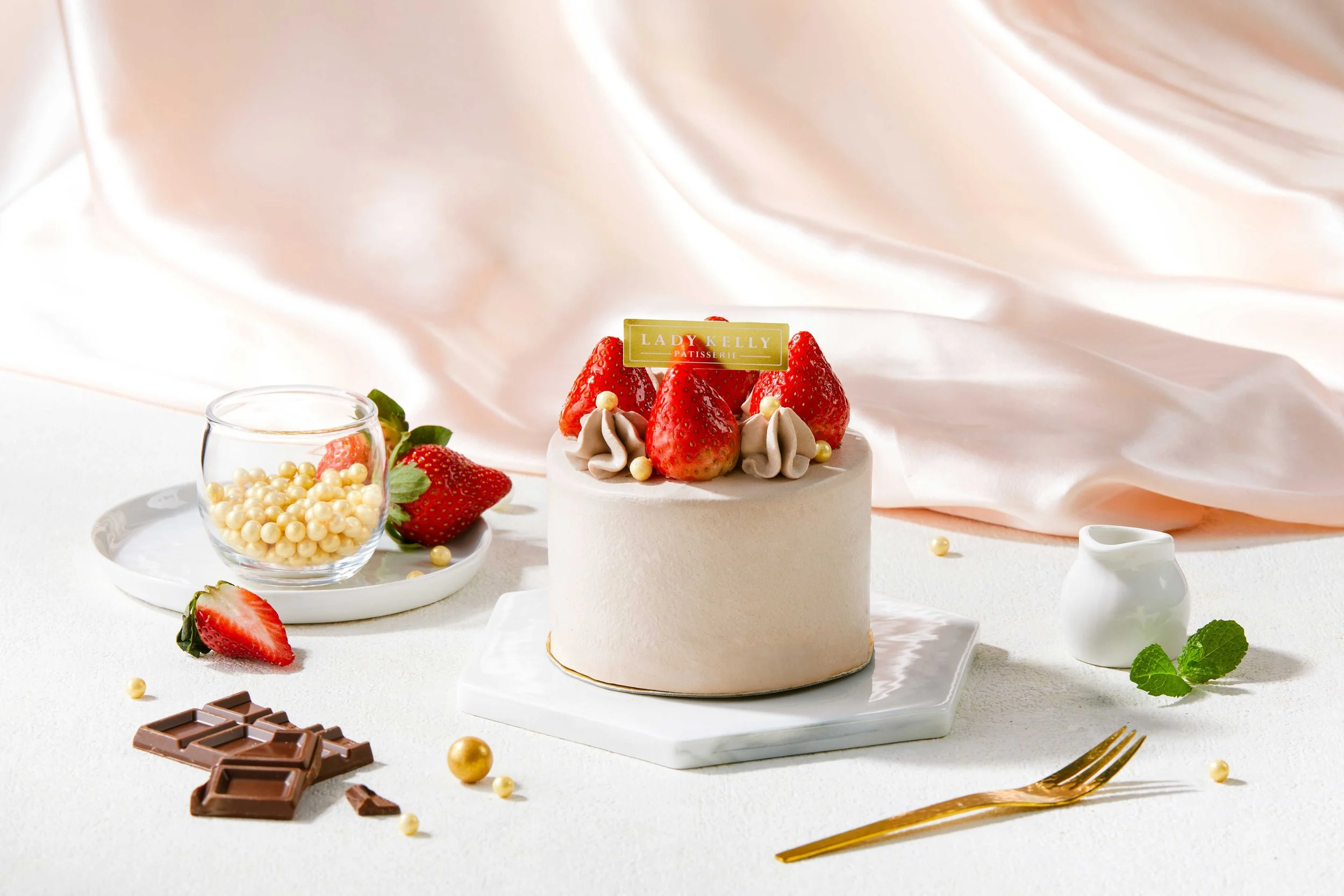 A round, white cake decorated with strawberries and small white pearls, with a gold label that says "Lady Kelly Patisserie" on top. Surrounding the cake are strawberries, a glass bowl of white pearls, a broken chocolate bar, some scattered gold pearl