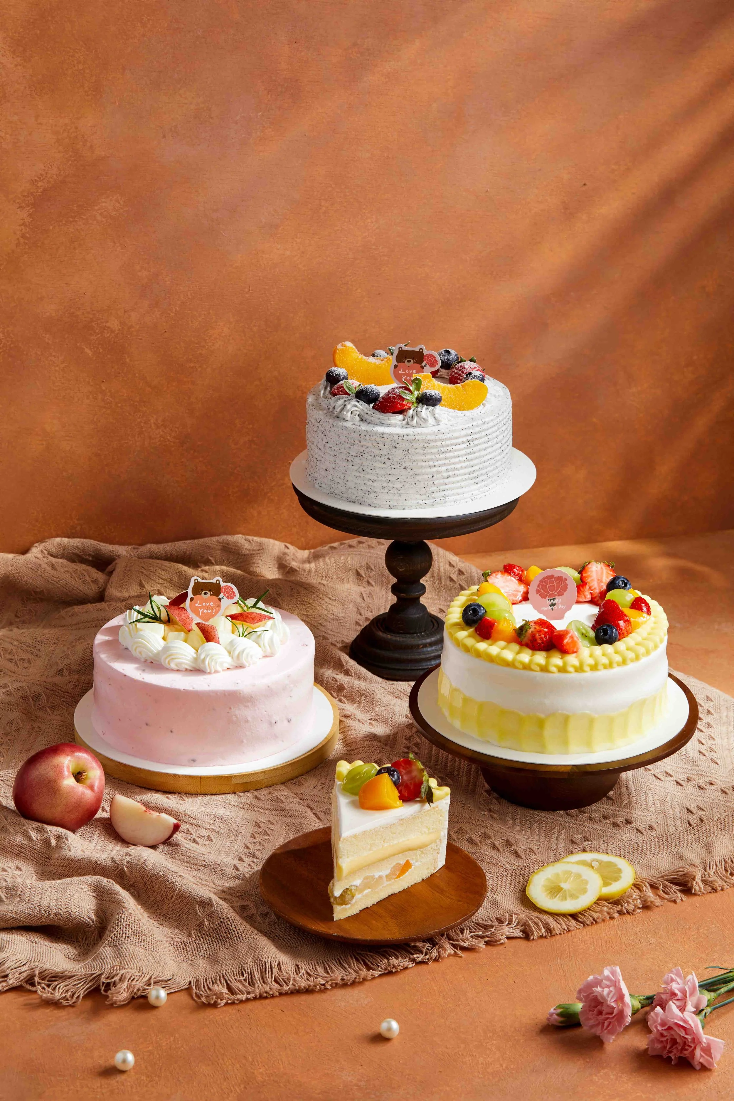 Assorted decorated cakes on a table with fruit toppings and floral decorations, along with an apple, lemon slices, and pink flowers nearby.