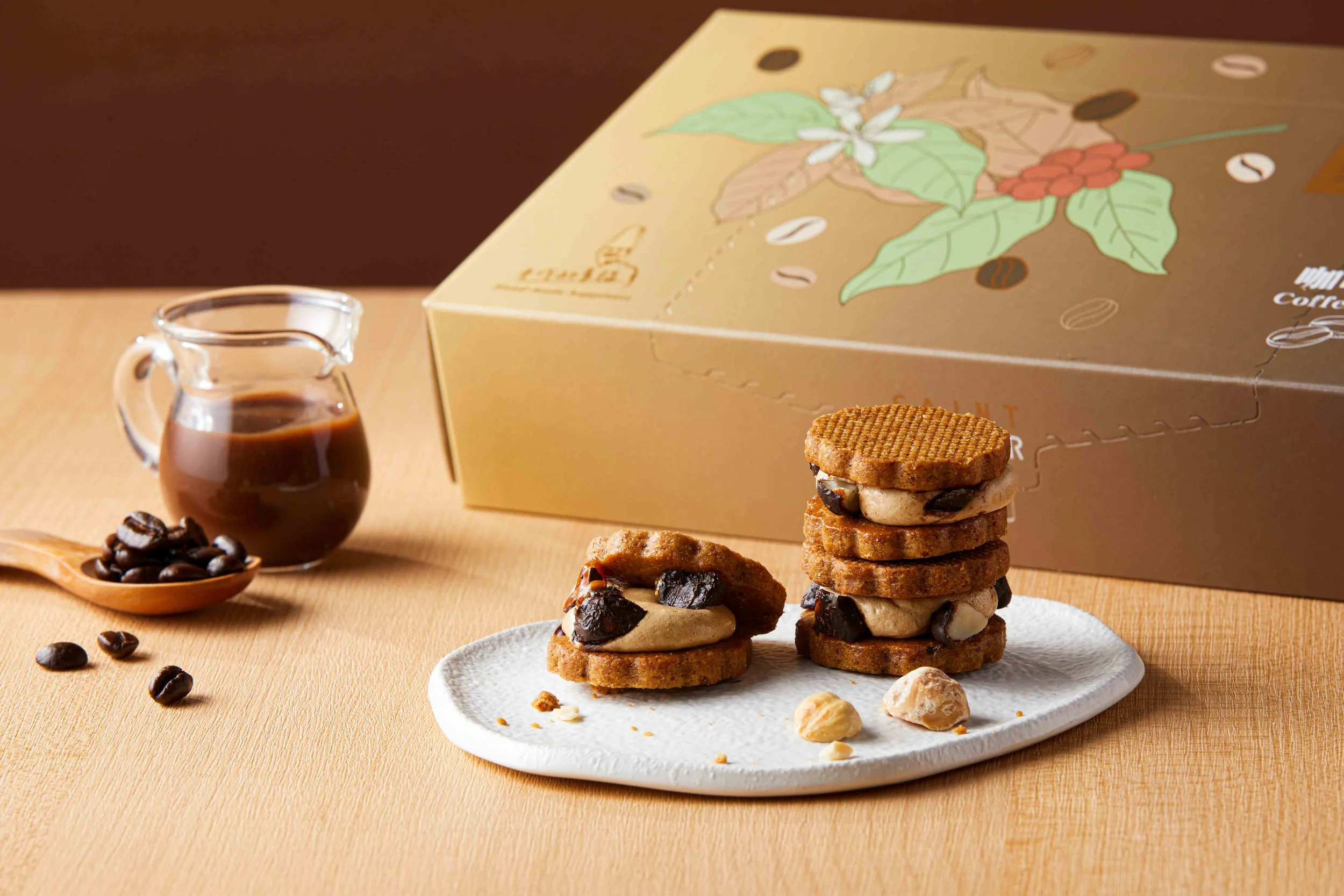 Stacked cookies with chocolate chips and nuts on a white plate, a small glass pitcher of coffee, and coffee beans scattered on a light wooden surface, with a partially visible box of coffee products in the background.