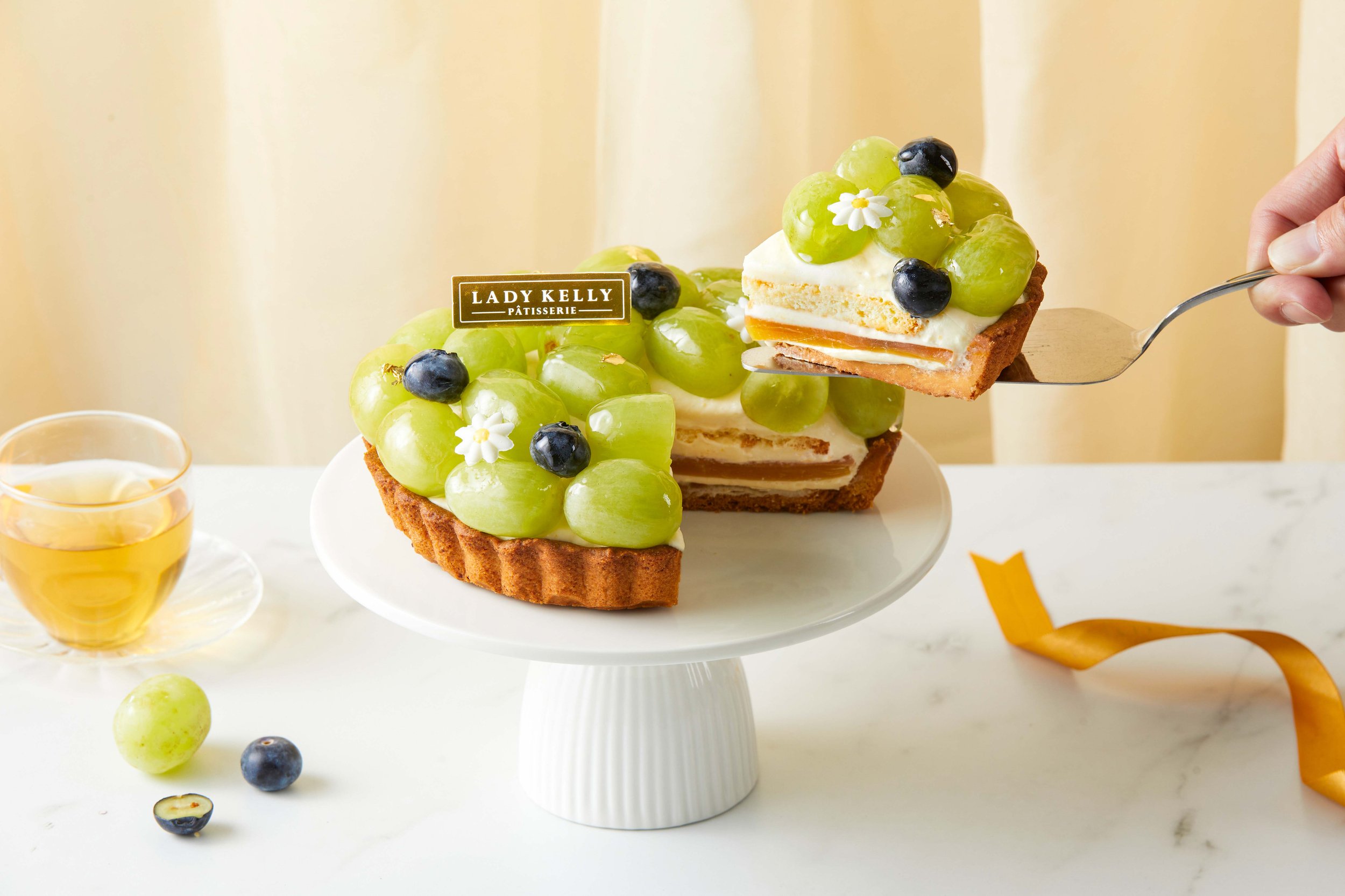 A slice of fruit tart with green grapes, blueberries, and small white flowers on top, with a visible lady Kelly pâtisserie label. The tart is on a white cake stand, with a glass of honey and some loose grapes and blueberries around it.