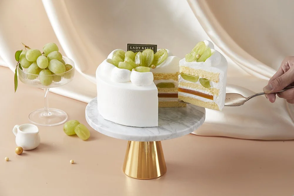 A white layered cake decorated with green grapes on top, placed on a marble cake stand, with a slice being served. There is a small glass bowl of green grapes next to the cake and a tiny white pitcher nearby.