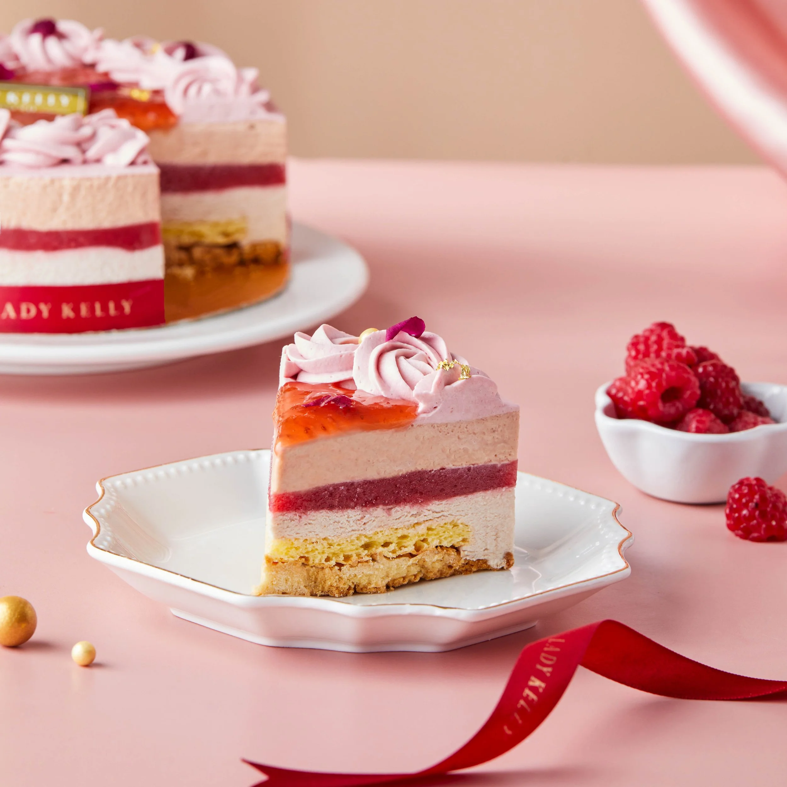 A multi-layered cake with pink and white icing, decorated with pink flowers and gold accents, placed on a decorative white cake stand with a scalloped edge, with raspberries and gold-colored candies on a pink surface nearby.