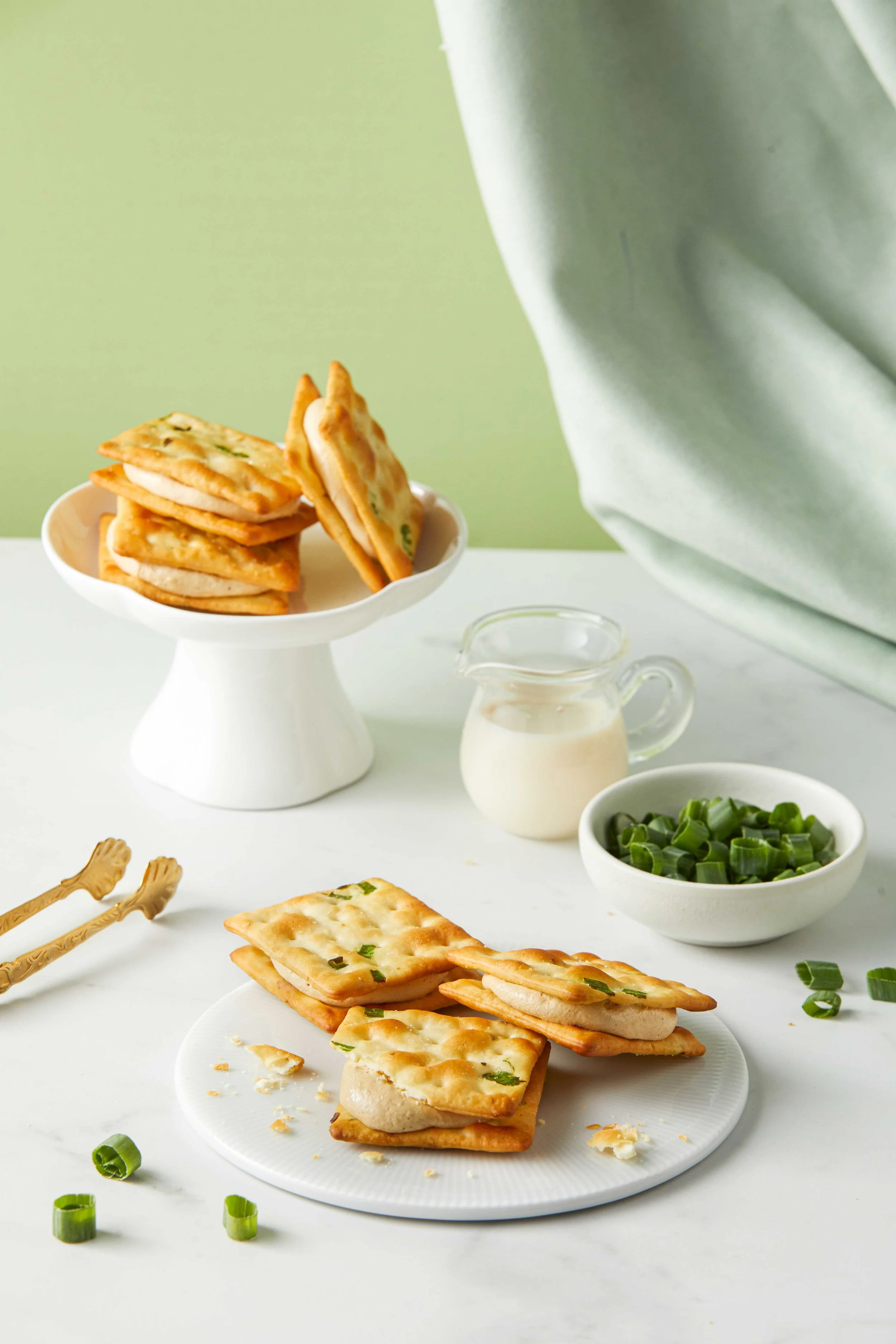 Plate of square-shaped crackers with cheese and chopped green onions, more crackers on a white surface, small bowl of chopped green onions, a pitcher of creamy dressing, and a bowl of green onions with a fork and a fork spoon on a white table, with a